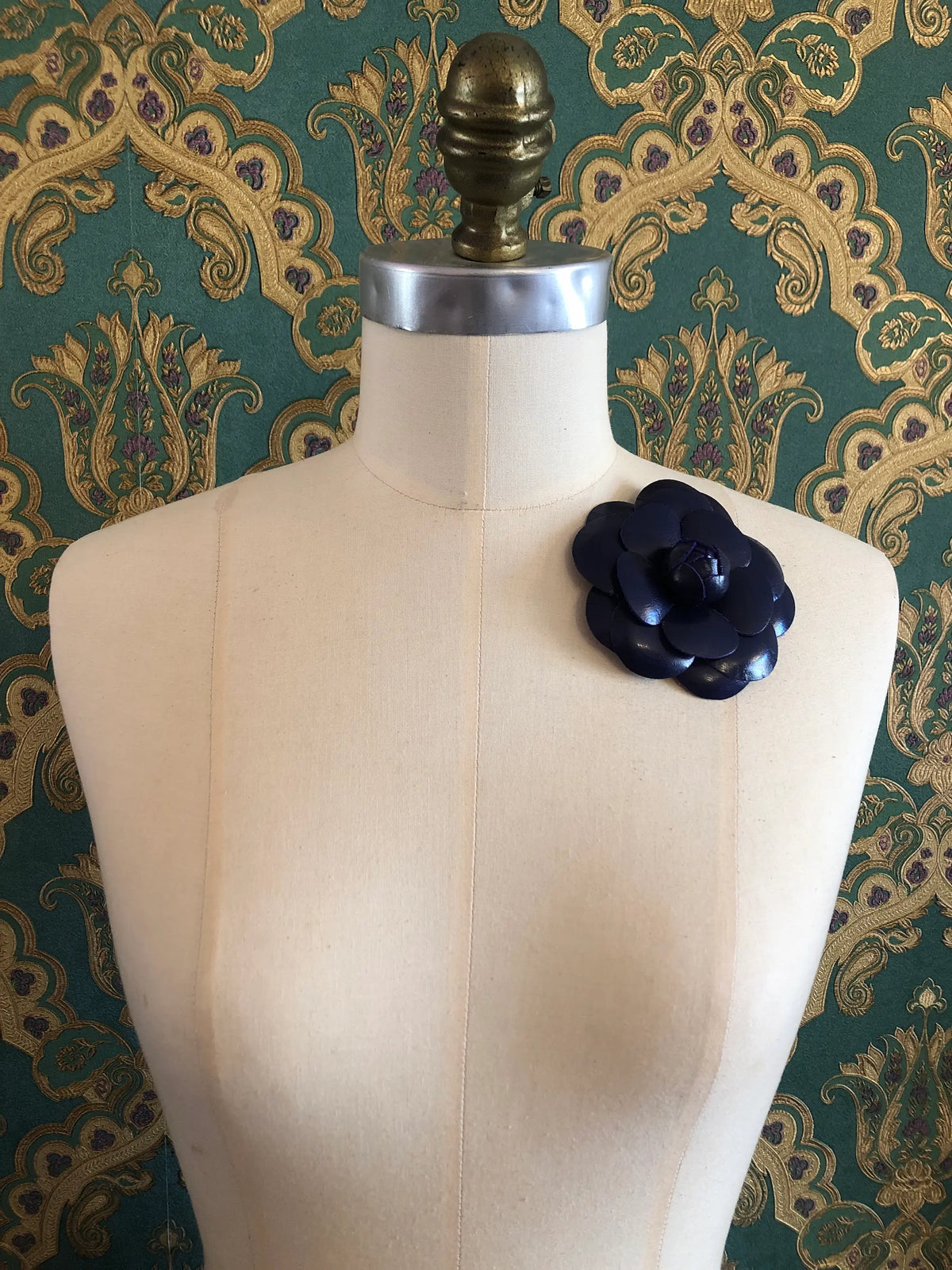 A Broochella Camelia dark purple leather  flower brooch displayed on a cream-coloured dress form against a green and gold damask patterned wallpaper background.