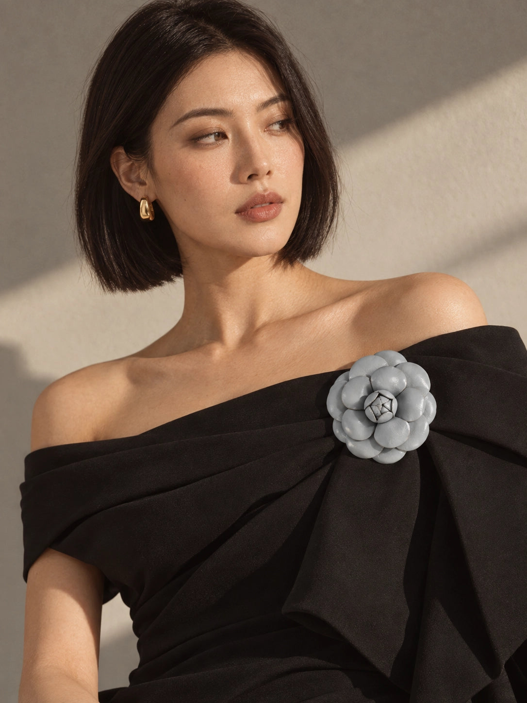 A woman with a short dark bob hairstyle wears an off-the-shoulder black dress featuring a Broochella Camelia  embellishment on the chest. She is photographed in warm, directional sunlight against a neutral beige wall.