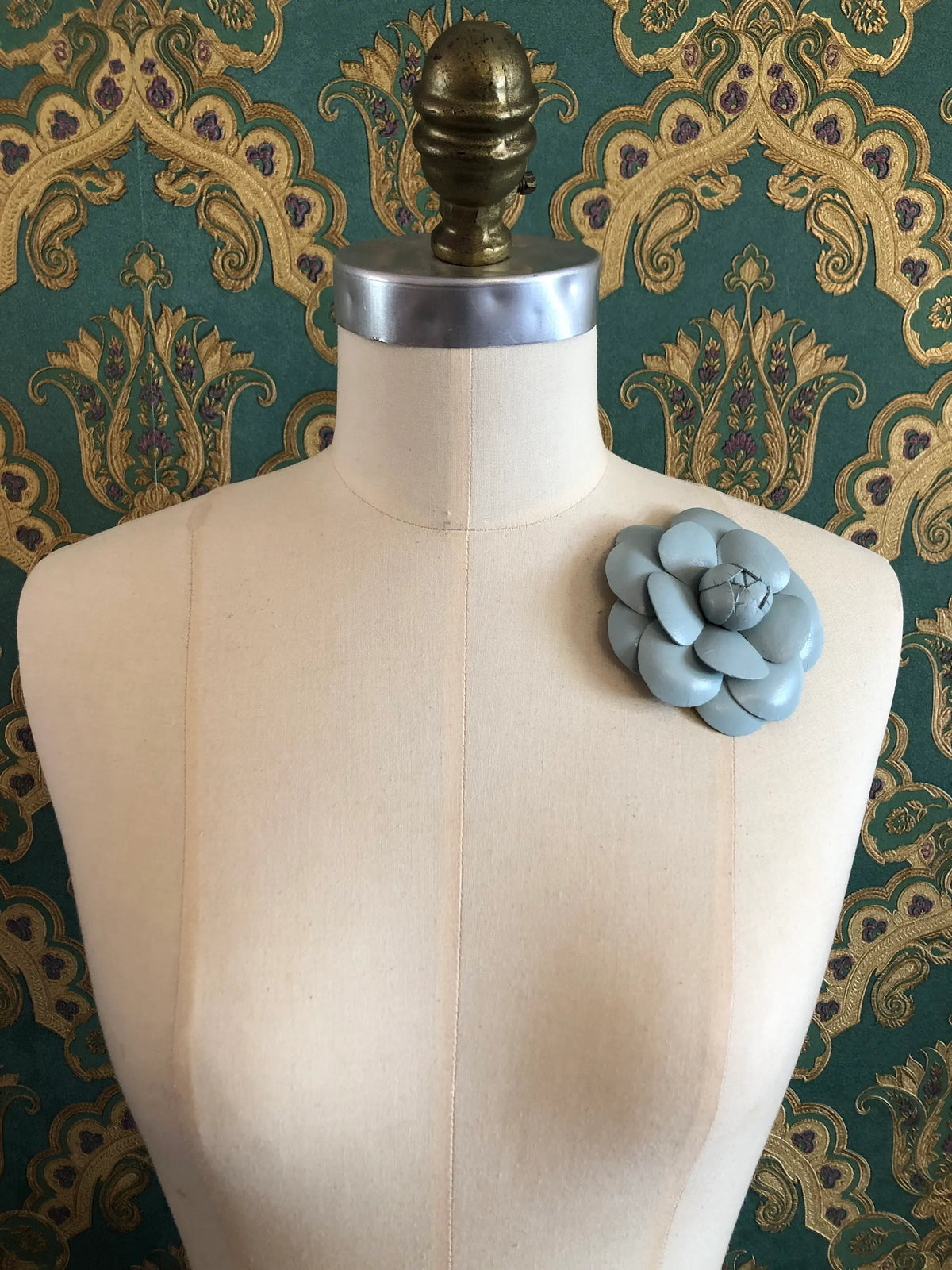 A Broochella Camelia mint leather flower brooch displayed on a cream-coloured dress form, set against a dark green damask patterned wallpaper background.