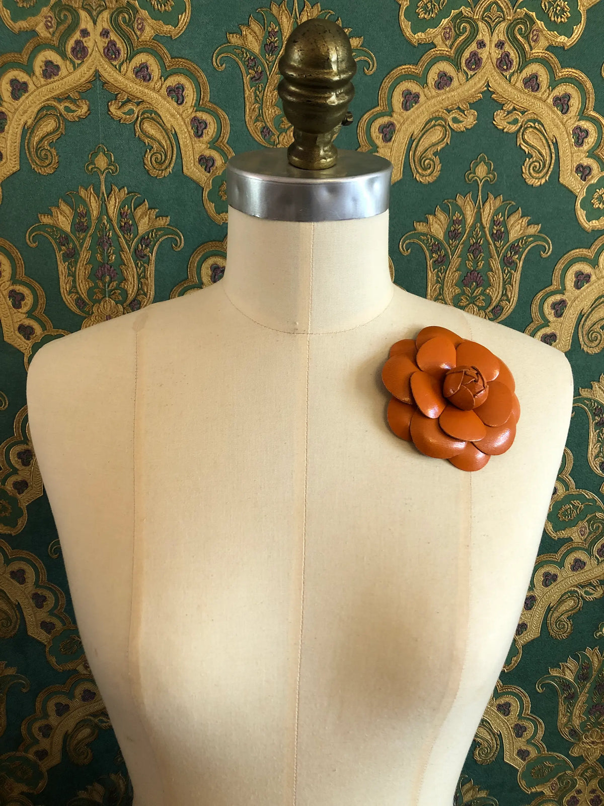 A Broochella Camelia orange leather flower brooch with layered petals is pinned to the left chest of a cream-coloured fabric dress form, set against a dark green and gold damask patterned wallpaper background.