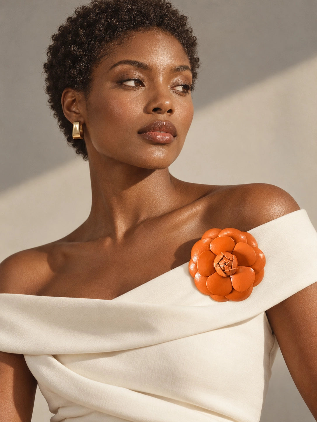 A close-up of a woman wearing a cream off-the-shoulder wrap dress with a Broochella Camelia handmade leather flower brooch embellishment on the shoulder, set against a neutral beige background with dramatic shadows.
