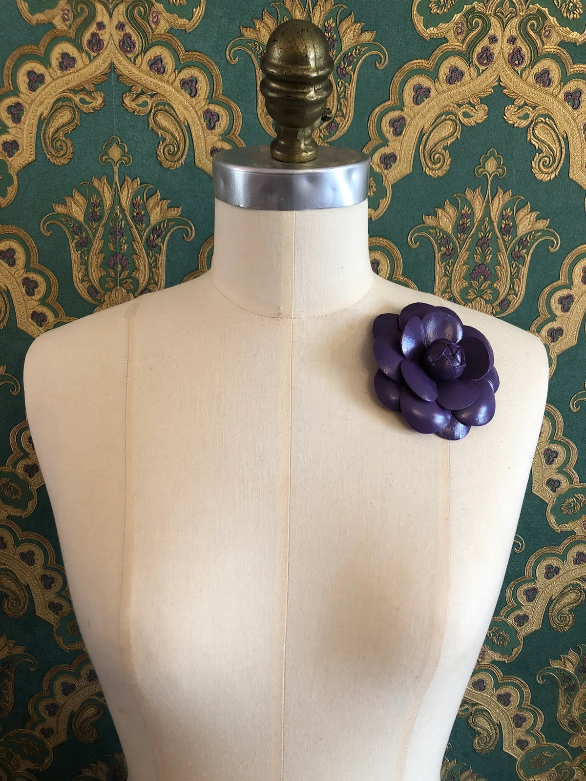 A Broochella Camelia purple leather flower brooch displayed on the left chest of a cream-coloured dress form mannequin, set against a green and gold damask patterned wallpaper background.