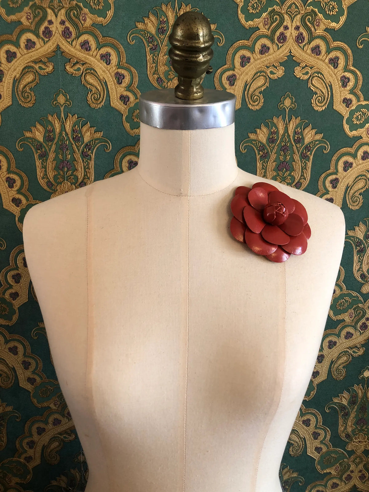 A Broochella Camelia red leather flower brooch displayed on a cream-coloured dress form against a green and gold damask patterned wallpaper background.