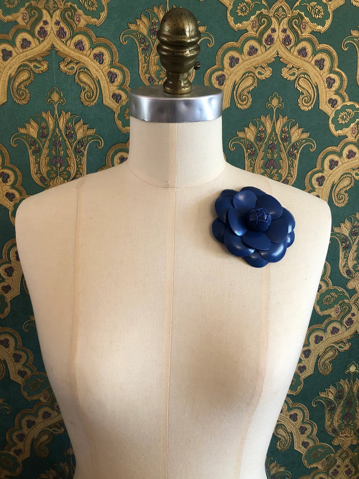 A Broochella royal blue fleather  flower brooch displayed on a cream-coloured dress form against a green and gold patterned wallpaper background.
