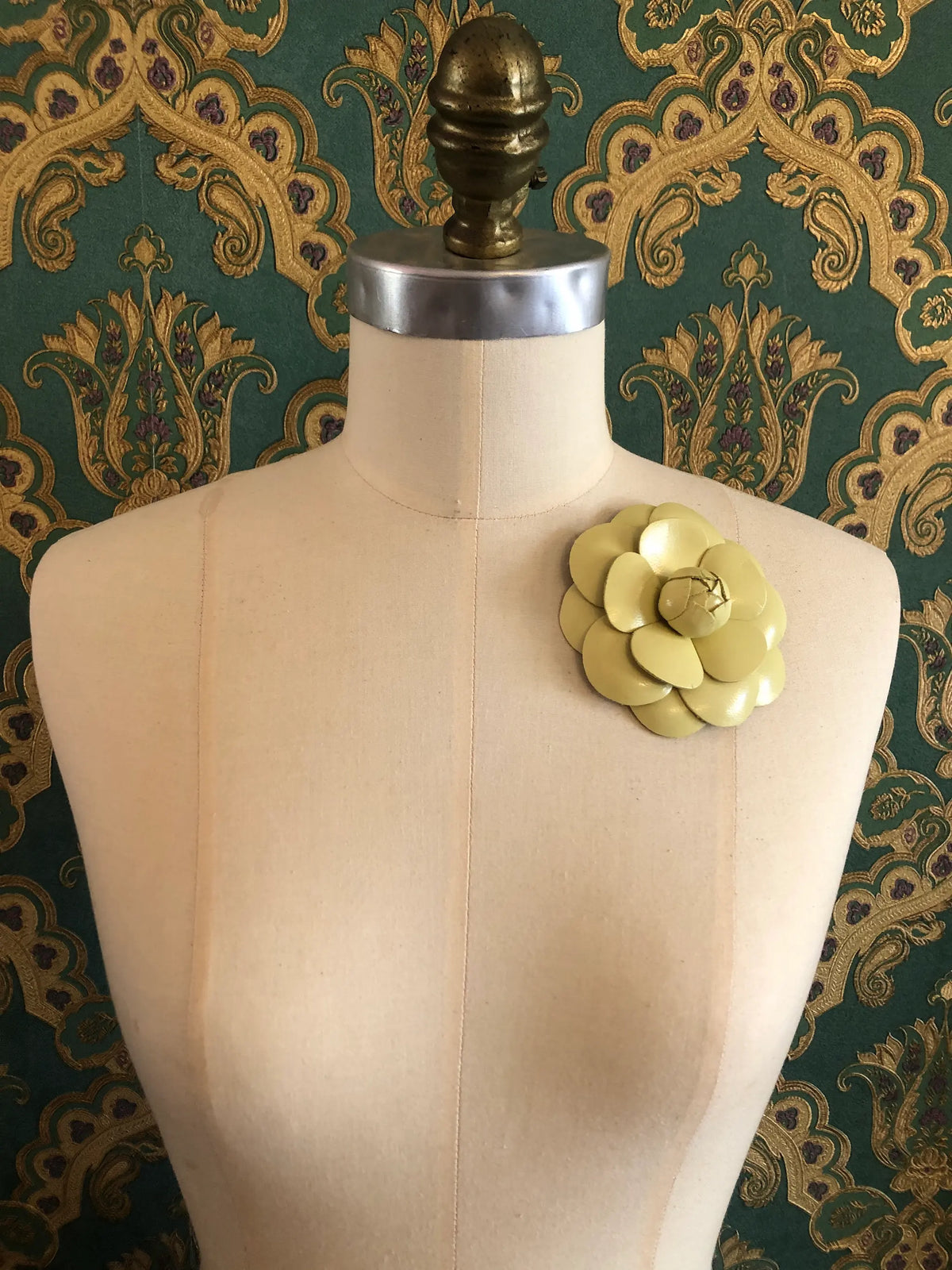 A  Broochella Camelia pale yellow fabric leather brooch pinned to the chest of a beige dress form, set against a dark green and gold damask patterned wallpaper background.