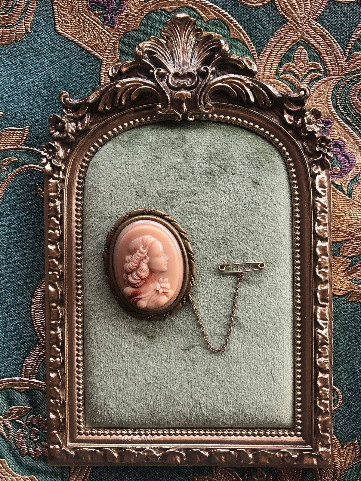 Broochella Cameo della Contessa Vintage Brooch Pin featuring a blush pink cameo portrait in an ornate antique gold-tone setting with safety chain, displayed on sage green velvet in an ornate gold frame against green and gold patterned fabric.