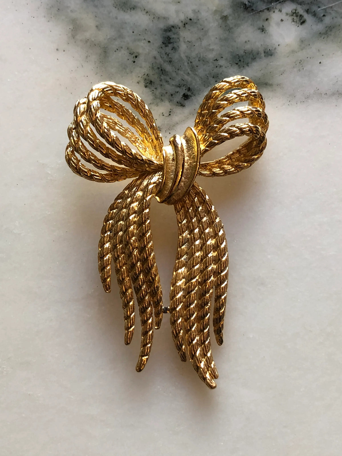 Broochella Camilla Vintage Gold Rope Bow Brooch featuring twisted gold-tone rope loops and long textured ribbon ends, photographed on a cream and grey marble background.