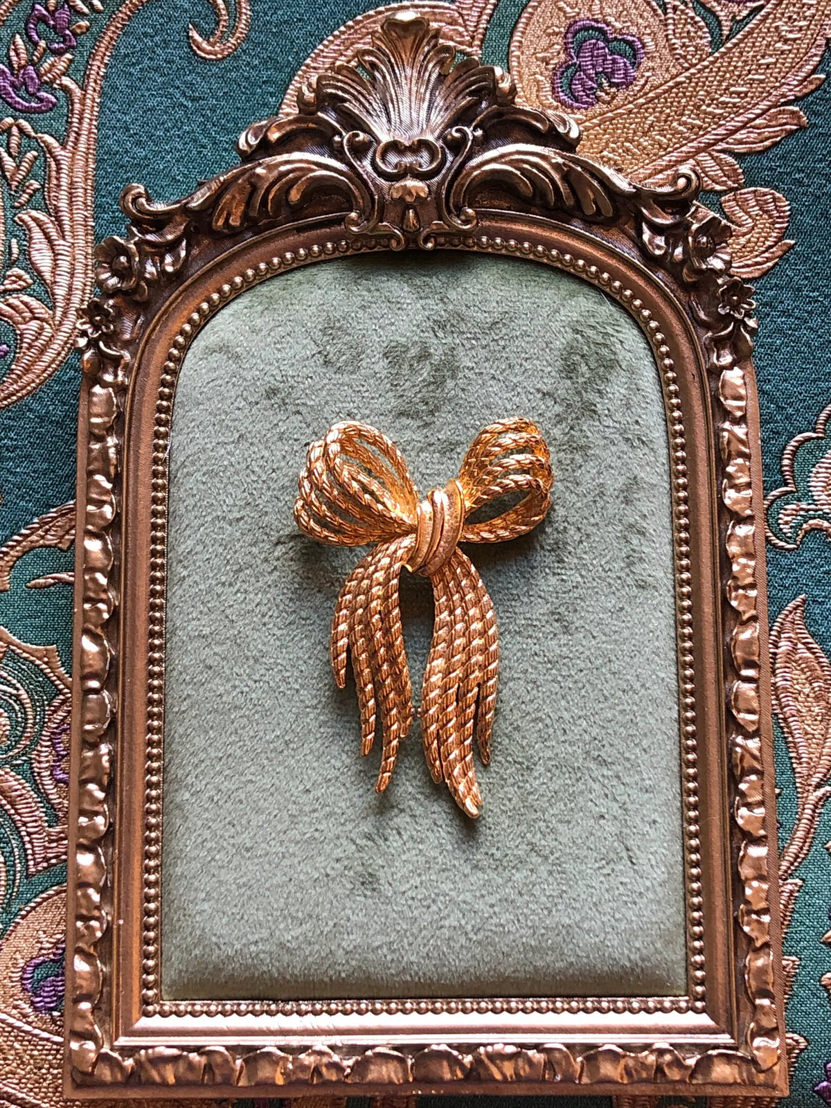 Broochella Camilla Vintage Gold Rope Bow Brooch featuring twisted gold-tone rope loops and long textured ribbon ends. It is presented on a green velvet tray with an ornate gold frame. The background is green and gold damask wallpaper