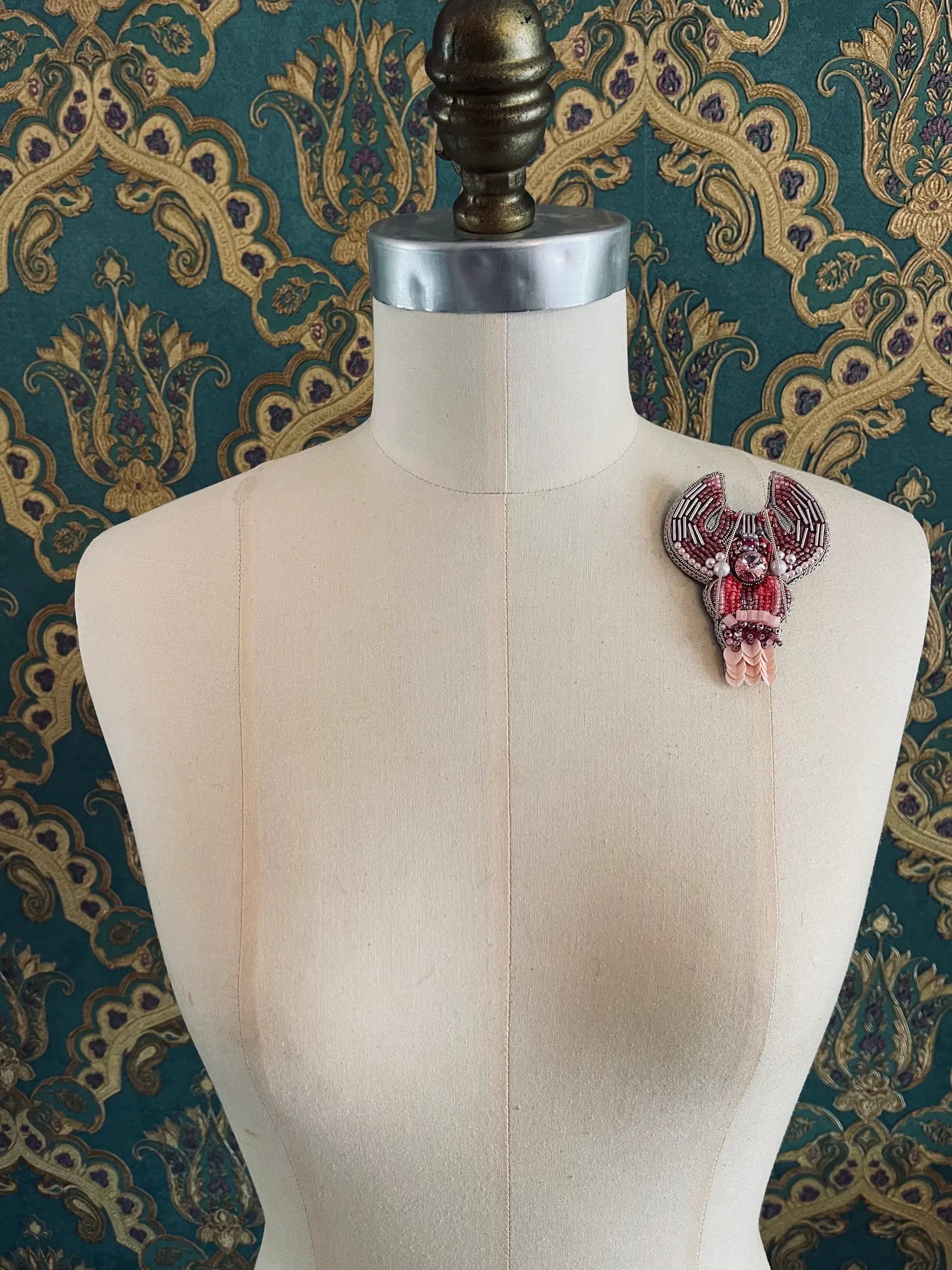 Broochella Cancer hand beaded pink silver designer zodiac brooch. Styled on a mannequin. The background is green and gold damask wallpaper