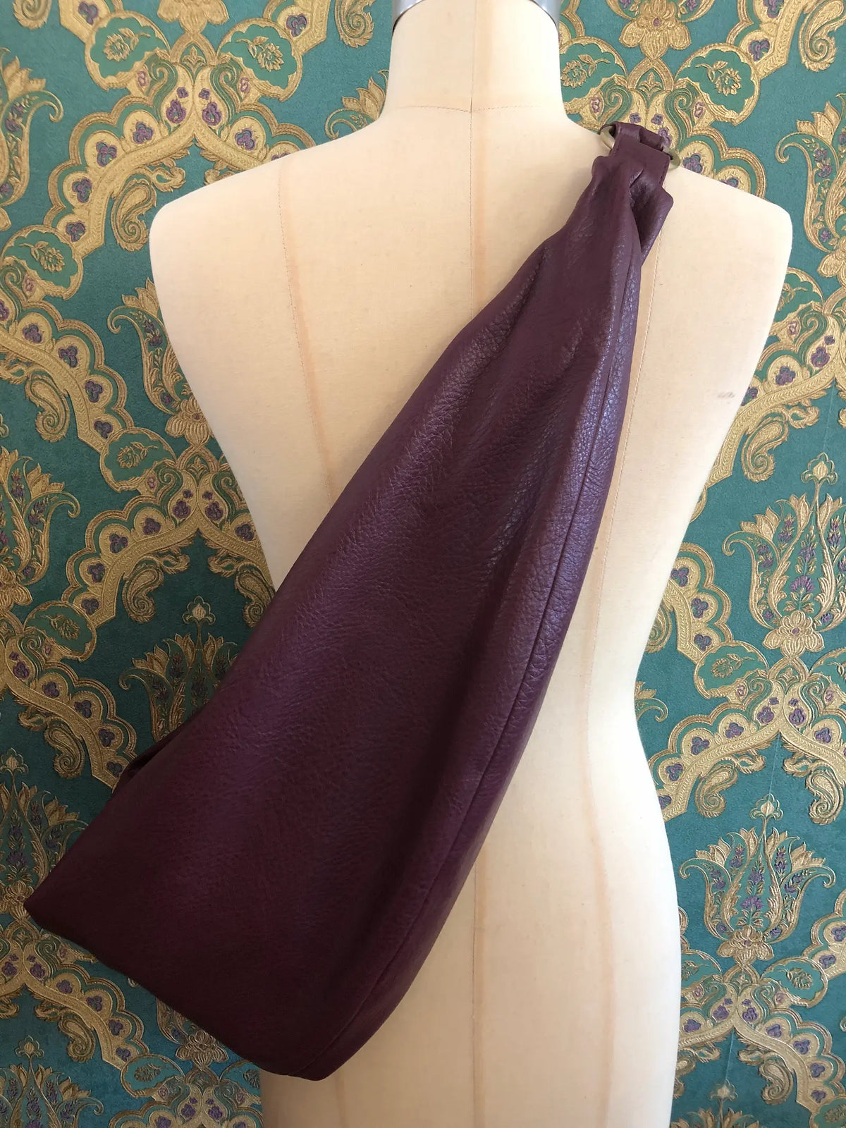 A Broochella Carla Rockmore merlot faux leather shoulder bag with a pebbled texture, displayed on a cream-coloured dress form mannequin against a green and gold damask patterned wallpaper background.