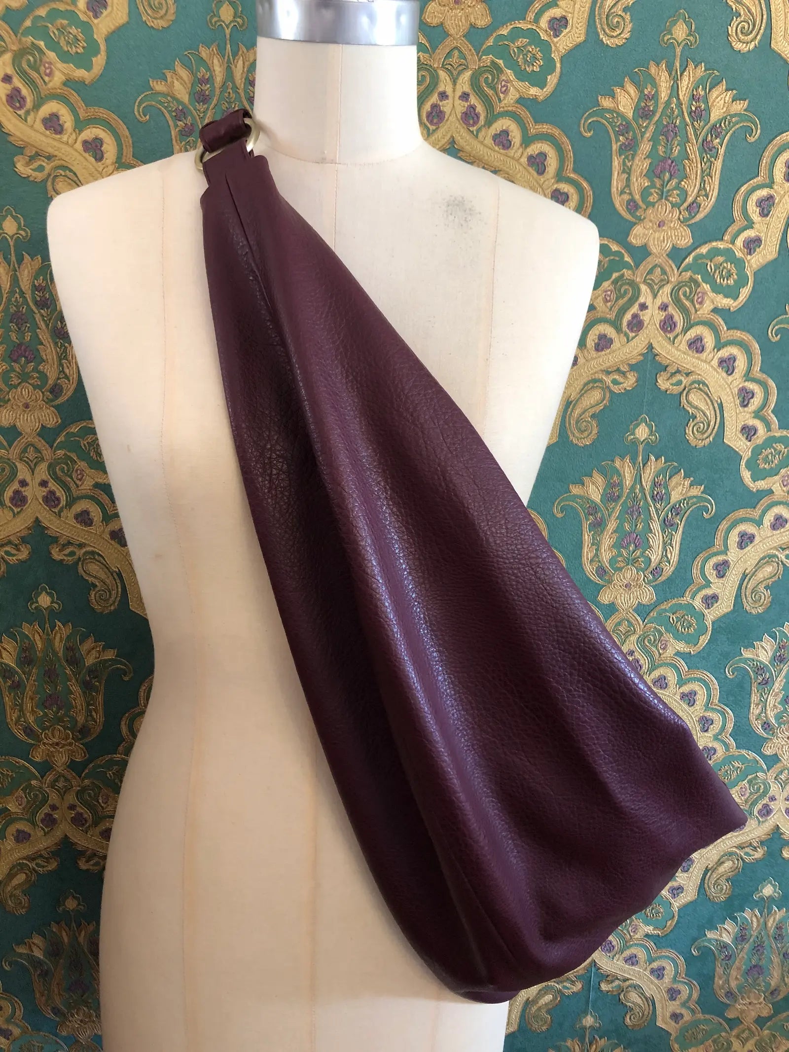A Broochella Carla Rockmore merlot faux leather shoulder bag with a pebbled texture, displayed on a cream-coloured dress form mannequin against a green and gold damask patterned wallpaper background.