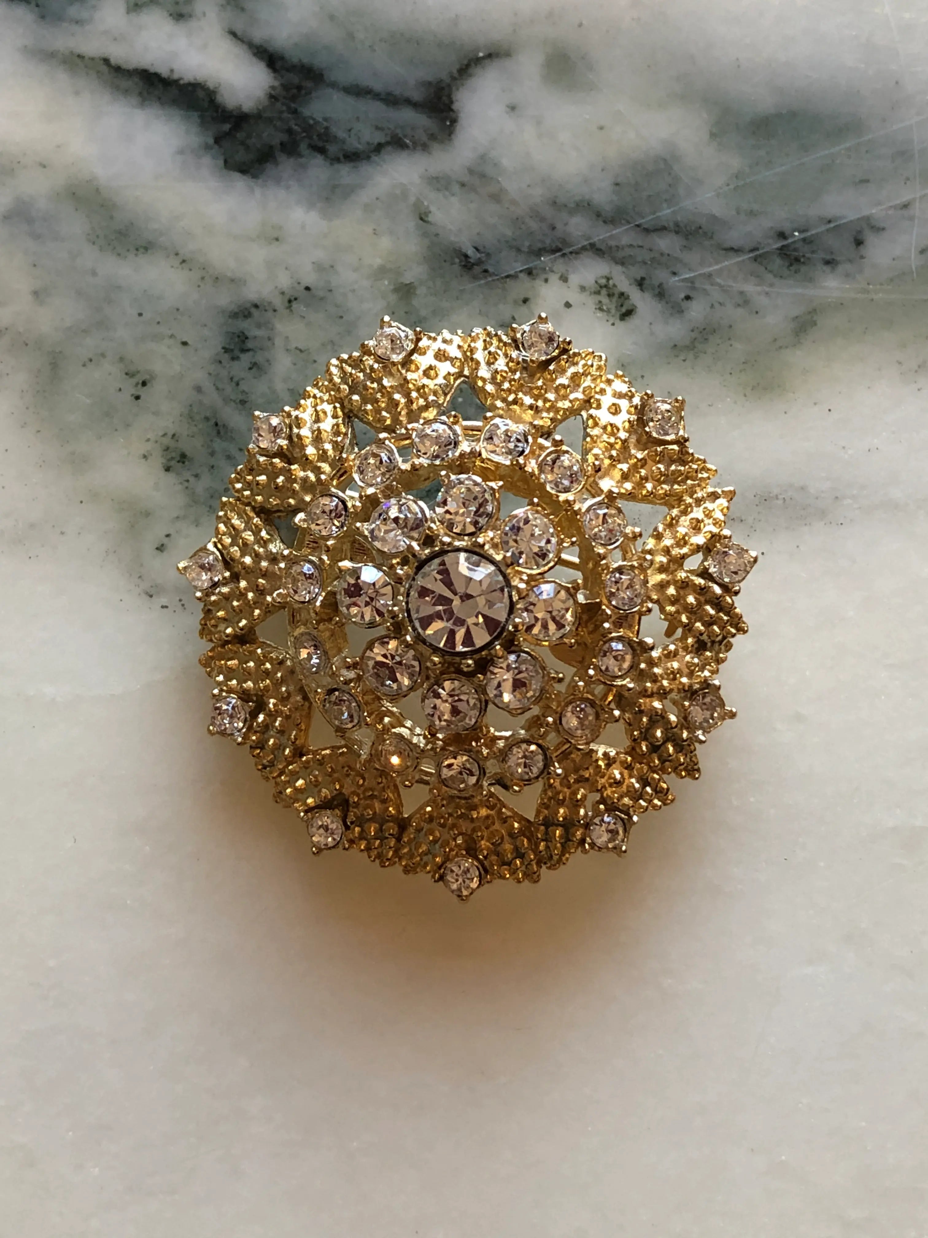 Broochella Caterina Vintage Gold Crystal Brooch featuring clear faceted crystals in an ornate textured gold-tone circular setting, photographed on a cream and grey marble background.
