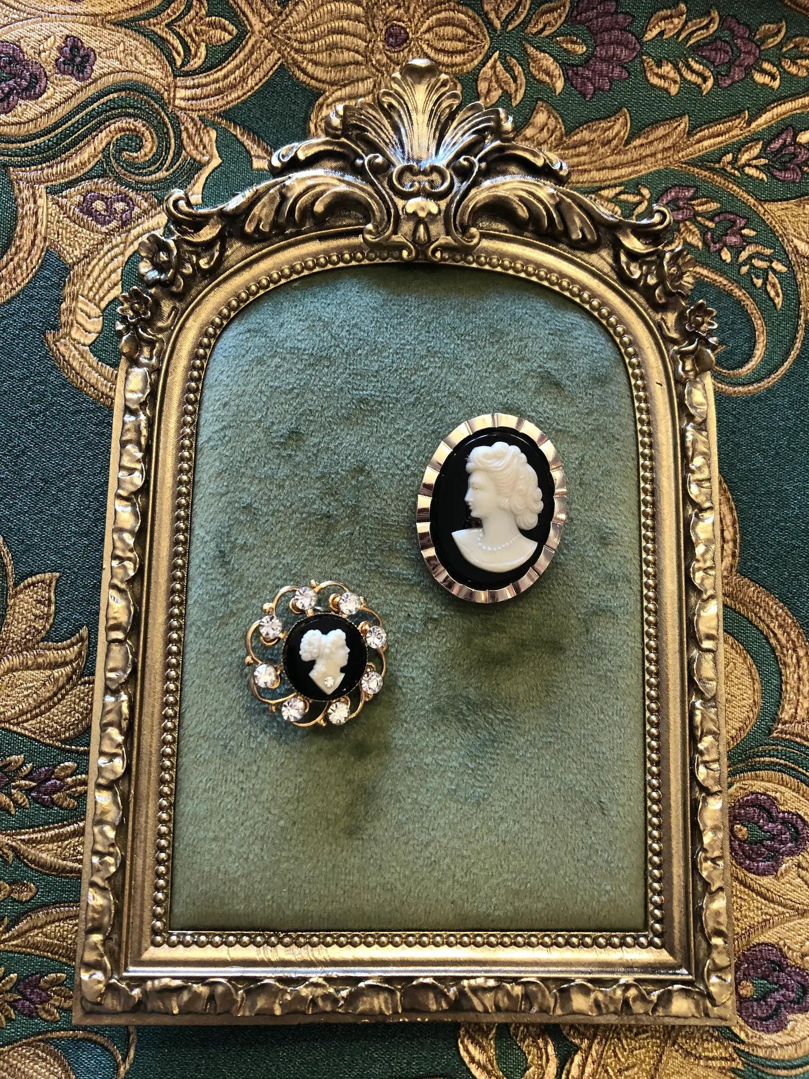 Broochella Cavaliere vintage cameo tie clip and brooch set  displayed on a green velvet insert inside an ornate gold frame, set against a patterned green and gold fabric background.