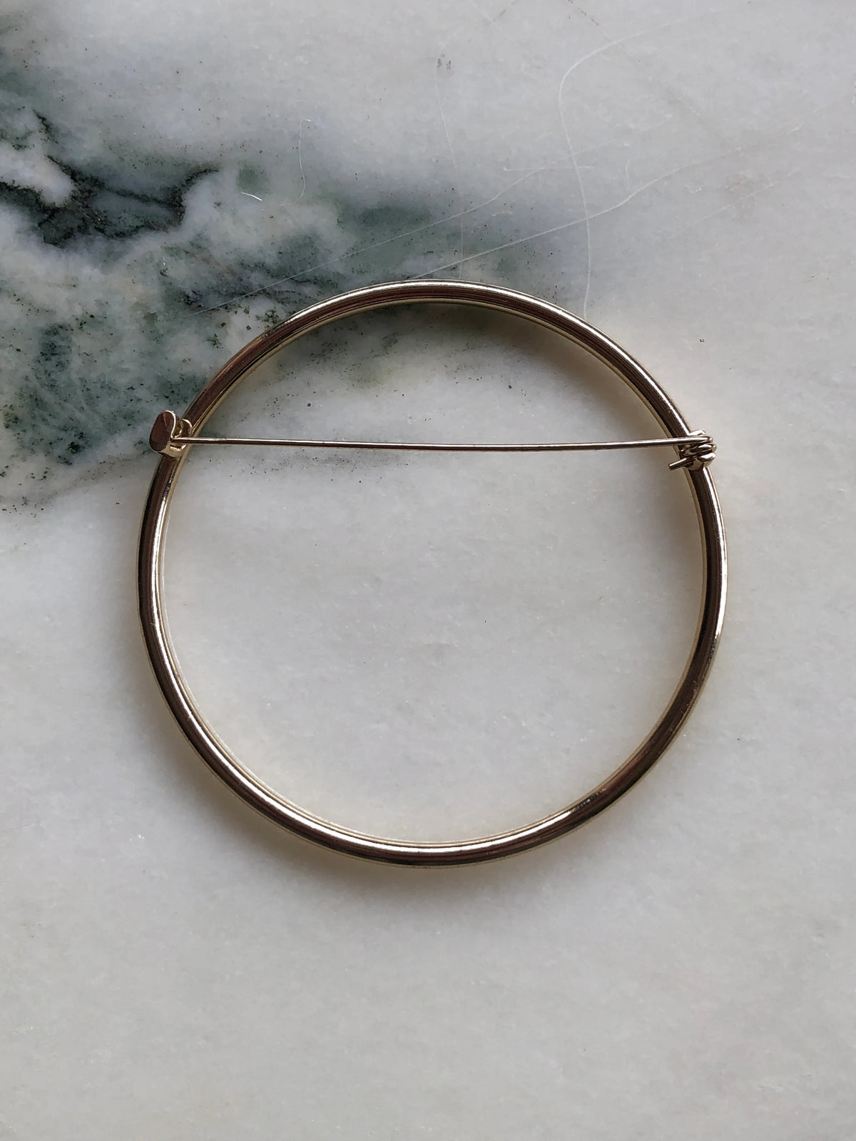Broochella Cercle de Paris Collectable Brooch back view showing the circular gold-tone metal design and traditional pin fastening, photographed on a white marble background with soft grey-green veining.