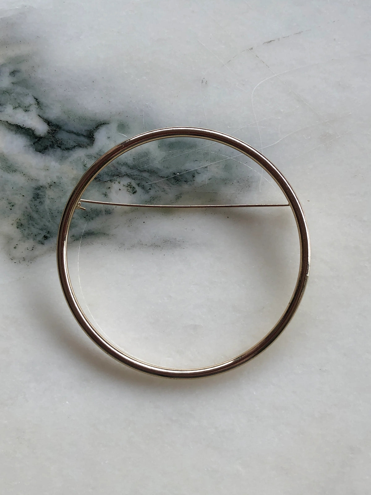 Broochella Cercle de Paris Collectable Brooch featuring a minimalist circular gold-tone metal design with a fine horizontal pin, photographed on a white marble background with soft grey-green veining.