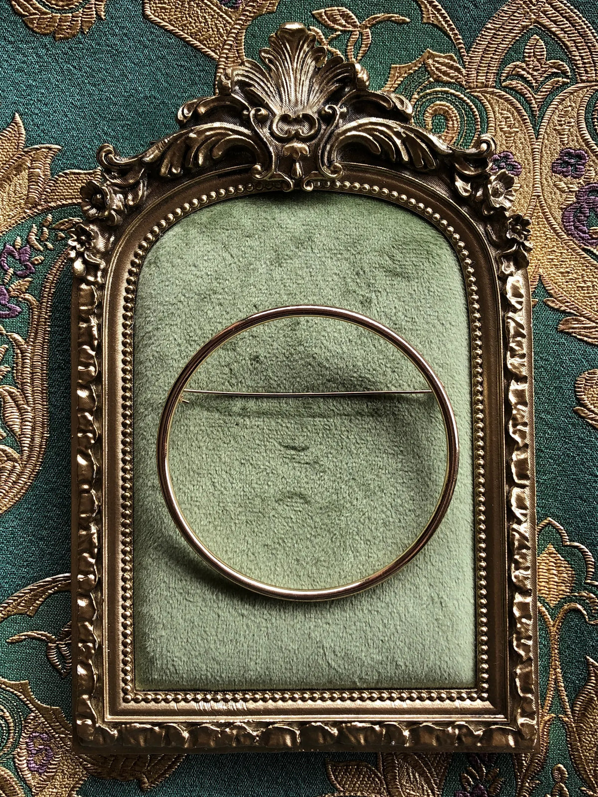 Broochella Cercle de Paris Collectable Brooch, a minimalist circular gold-tone brooch with a fine horizontal pin, displayed on sage green velvet in an ornate antique gold frame against green and gold patterned fabric.
