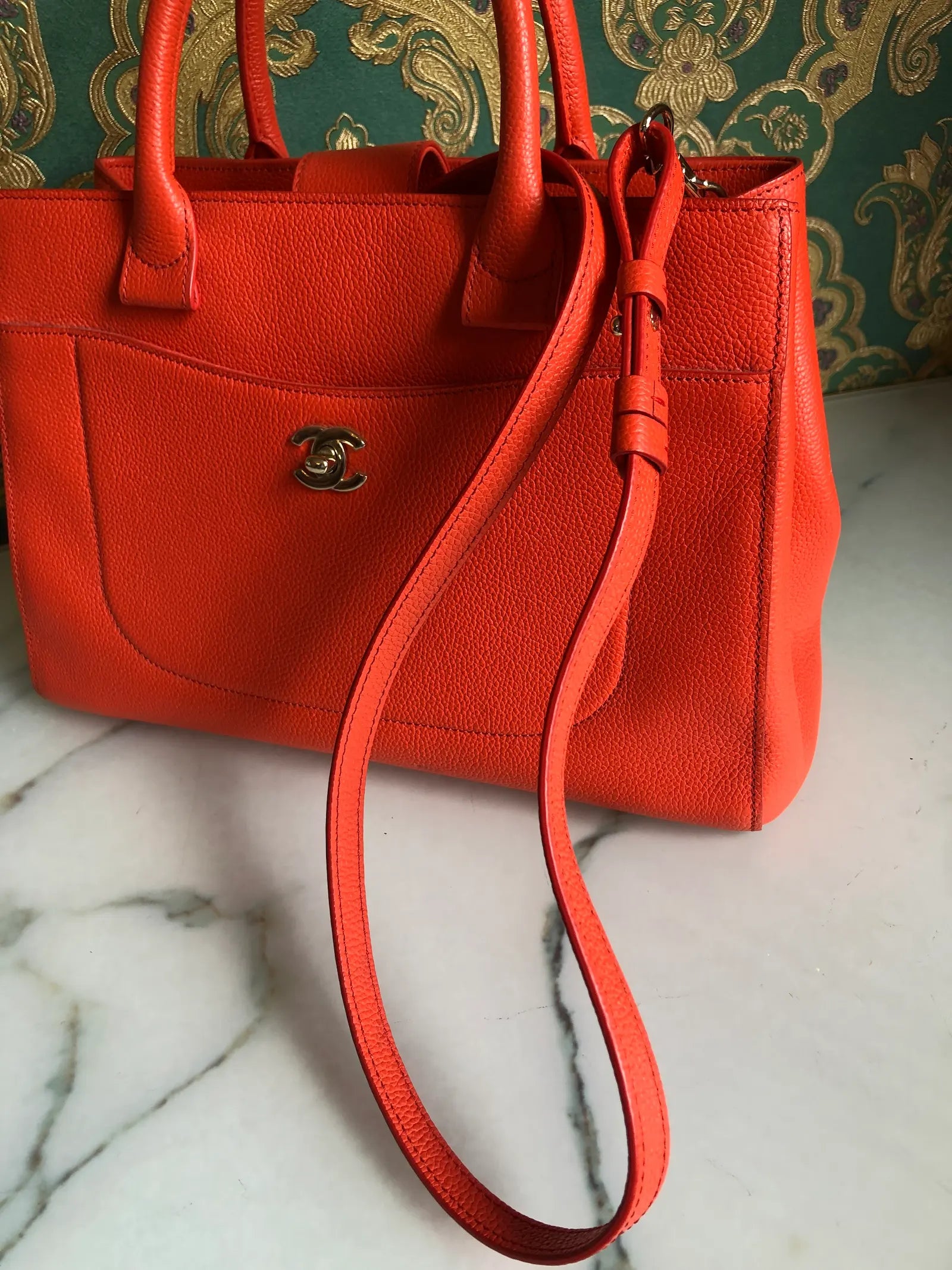 Broochella Pre-owned Chanel Executive Tote Bag in orange caviar leather with top handles and detachable shoulder strap. The background is green and gold damask wallpaper and a marble table