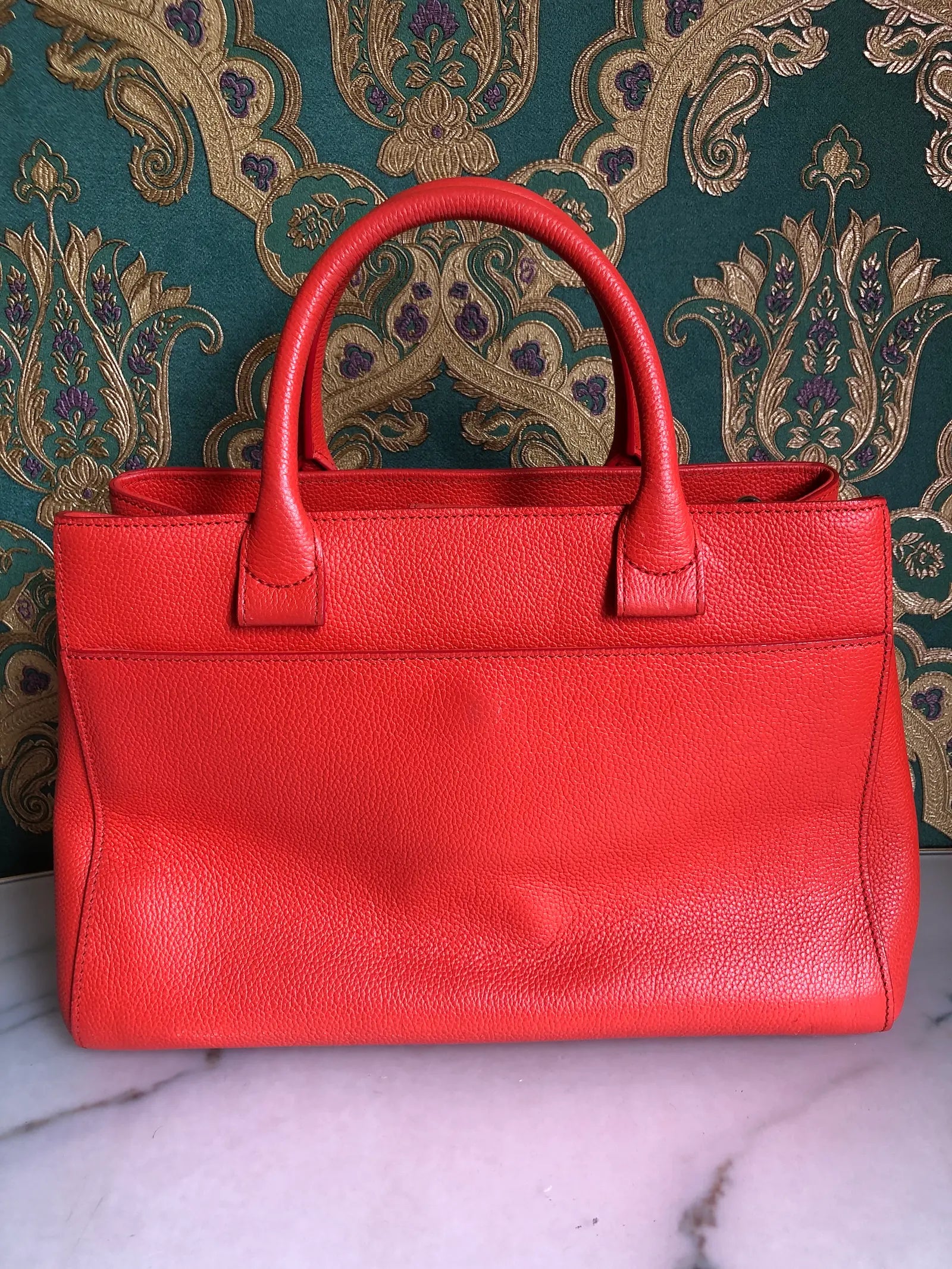 Broochella Pre-owned Chanel Executive Tote Bag in orange caviar leather with top handles and detachable shoulder strap. The background is green and gold damask wallpaper and a marble table