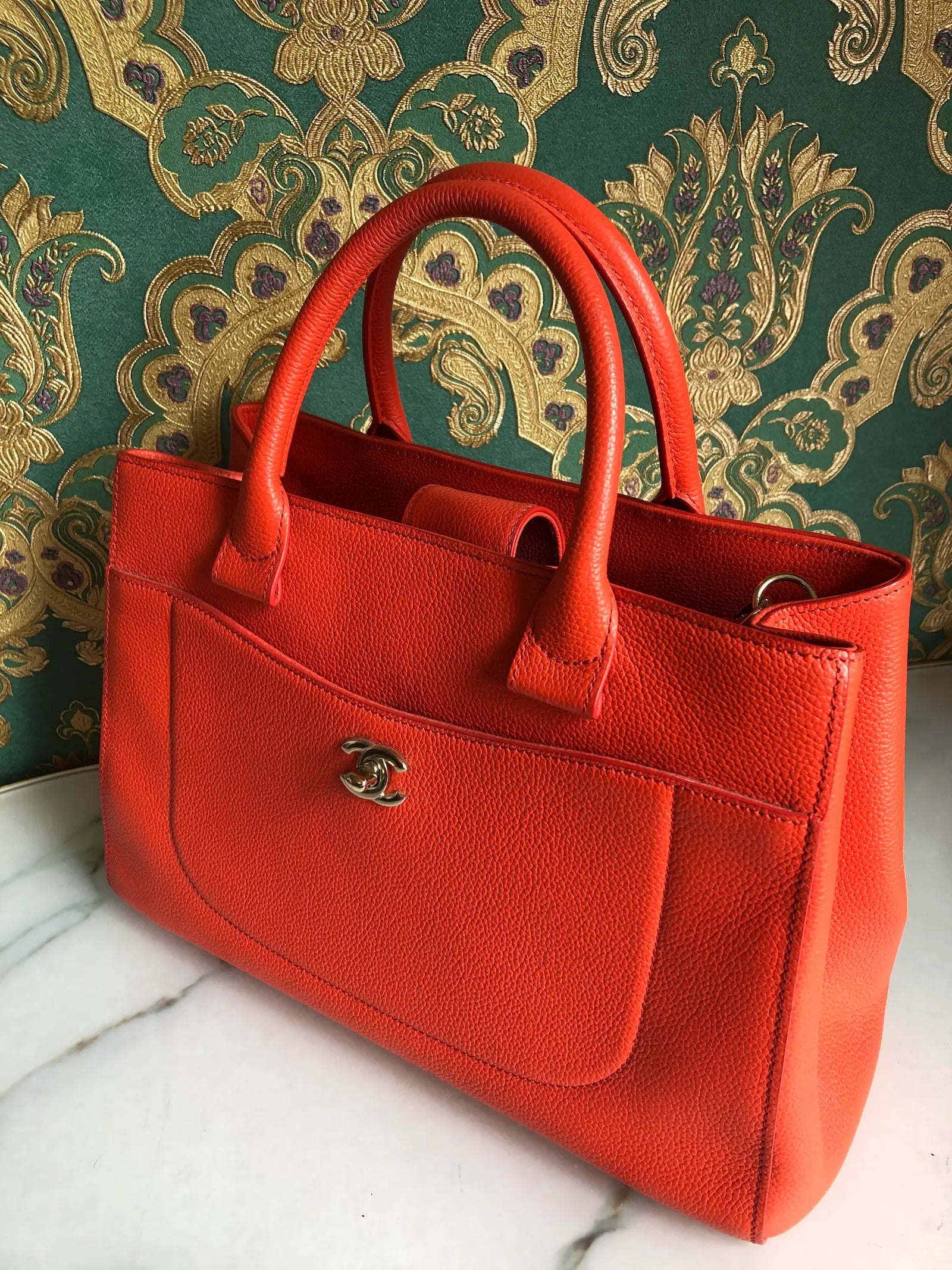 Broochella Pre-owned Chanel Executive Tote Bag in orange caviar leather with top handles and detachable shoulder strap. The background is green and gold damask wallpaper and a marble table