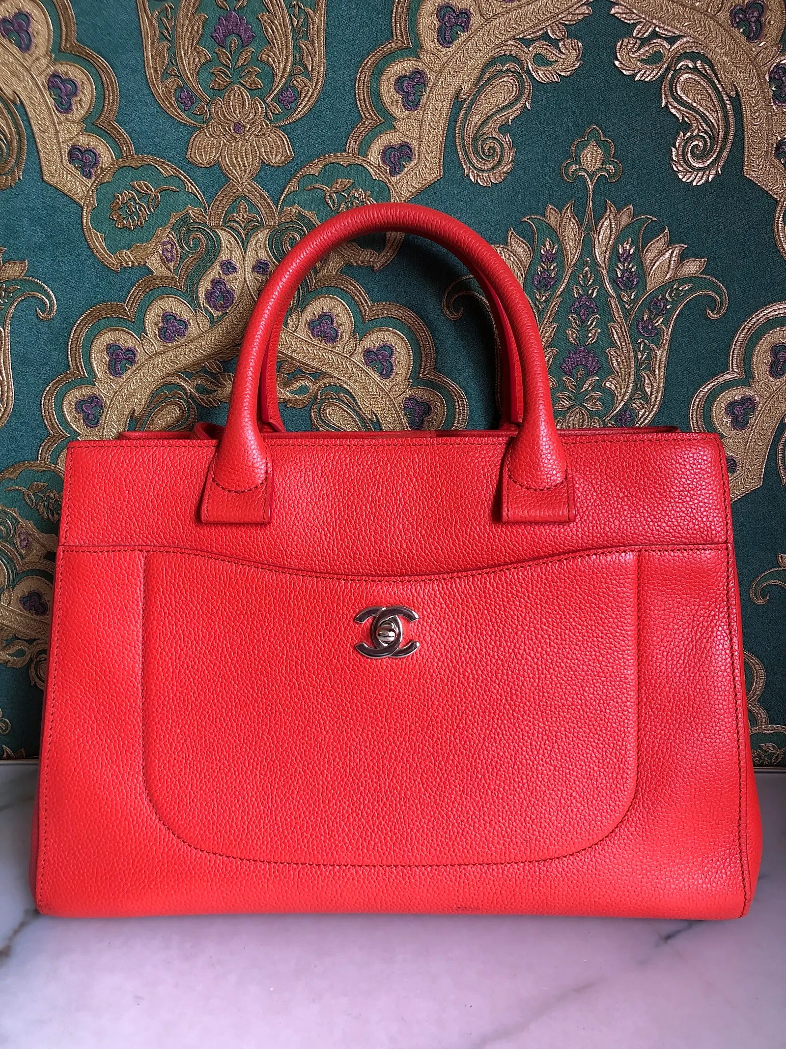 Broochella Pre-owned Chanel Executive Tote Bag in orange caviar leather with top handles and detachable shoulder strap. The background is green and gold damask wallpaper and a marble table