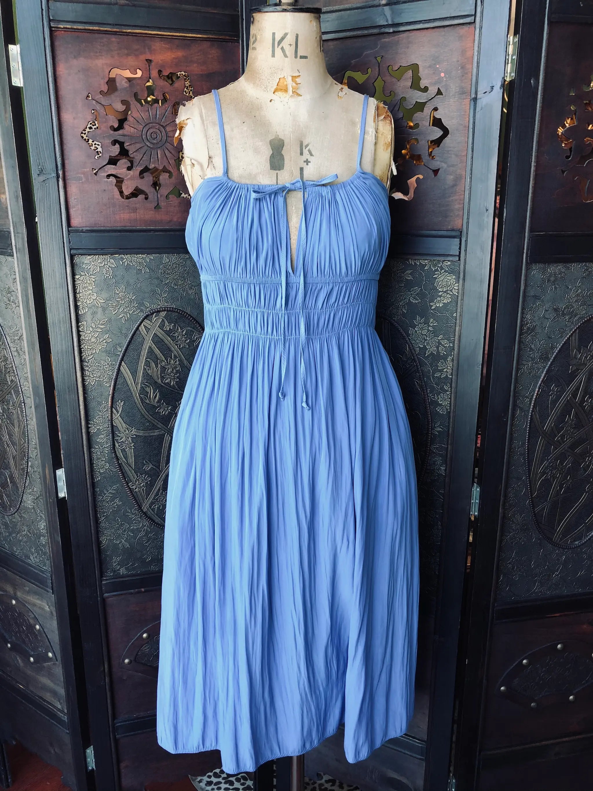 Broochella Ciao Lucia midi dress. Size medium, soft blue, 100% Polyester. I has spaghetti straps, gathered bodice with tie detail and an elasticated waist. It is pictured on a vintage mannequin. The background is an ornate, vintage, dark wood room divider. 