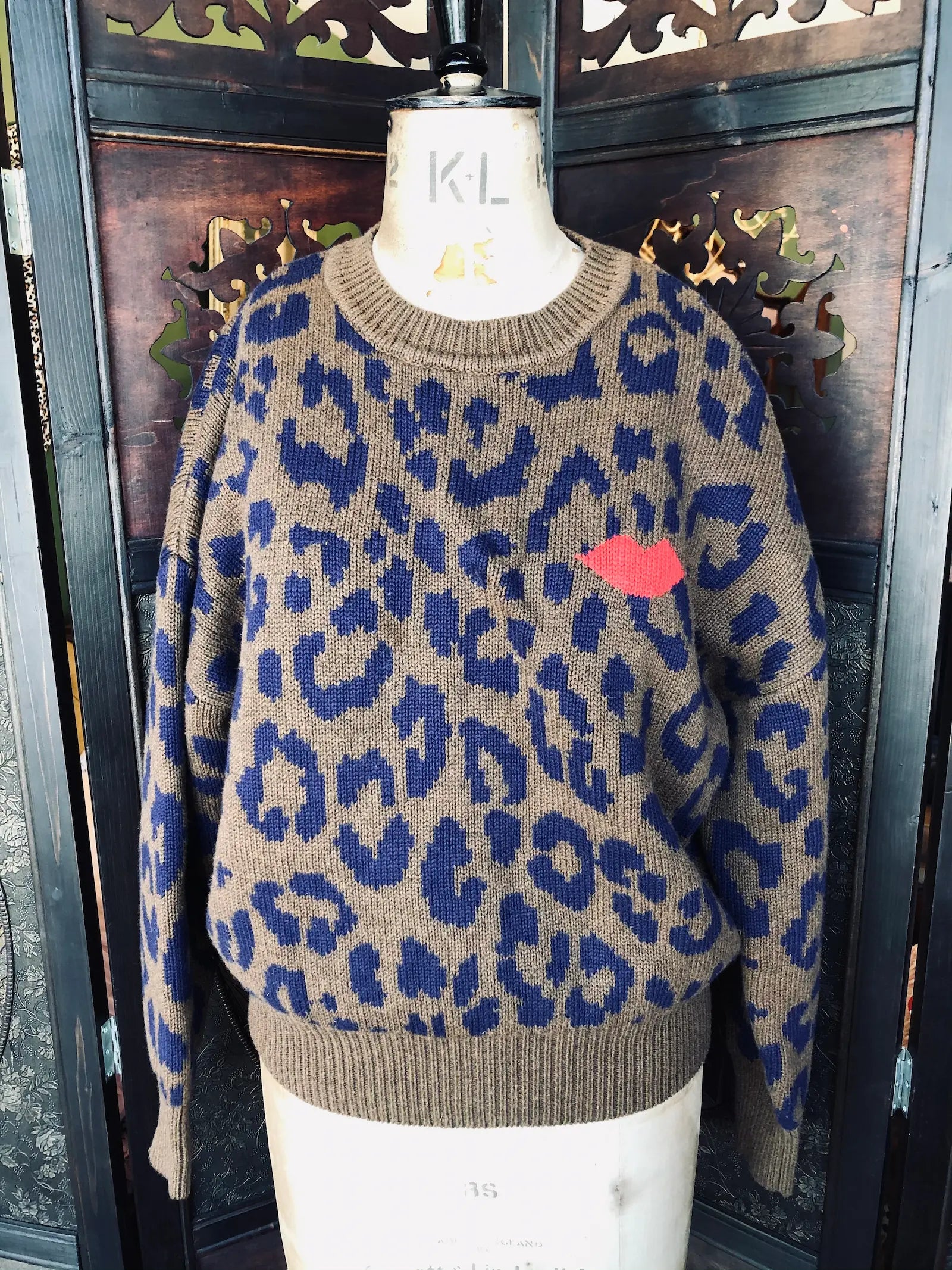 A Broochella Clare V knit crewneck sweater displayed on a mannequin torso, featuring a brown and blue leopard print pattern with a small red heart on the chest. The background consists of a dark wooden door with decorative cutouts.
