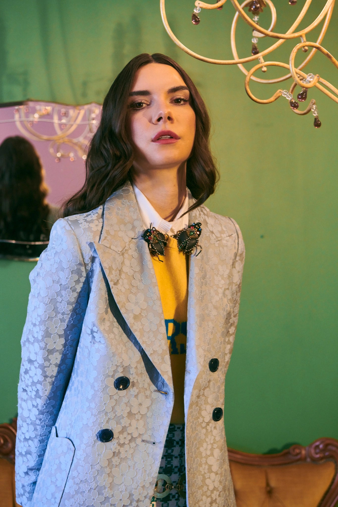 Dark haired female model wearing a pale blue floral blazer. She is wearing two Broochella cockroach brooches. The background is green. 