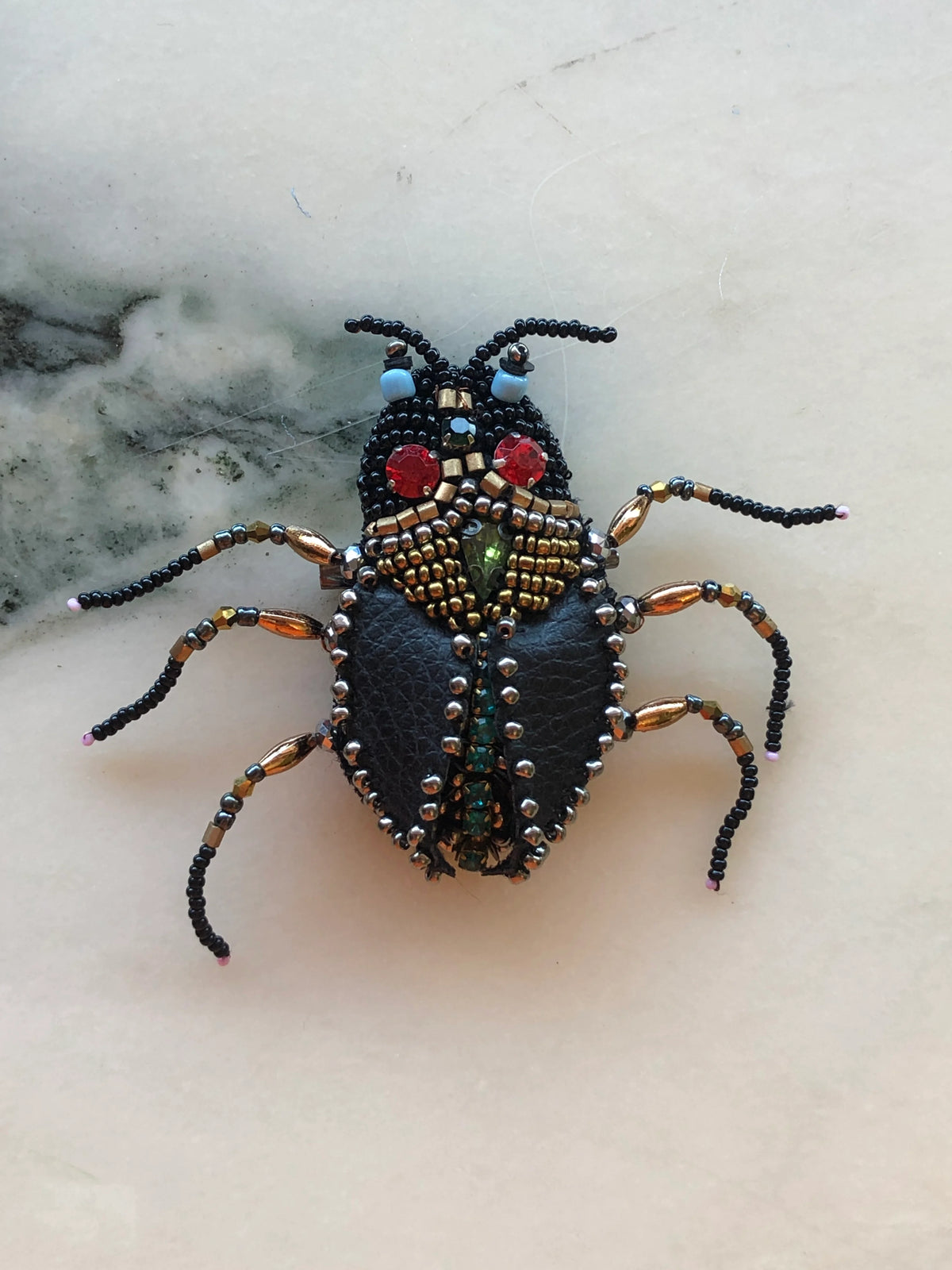 Broochella Cockroach Brooch Pin featuring a sculptural insect design hand embellished with black, gold and metallic beads, red crystal eyes and textured black wings on a cream marble background.