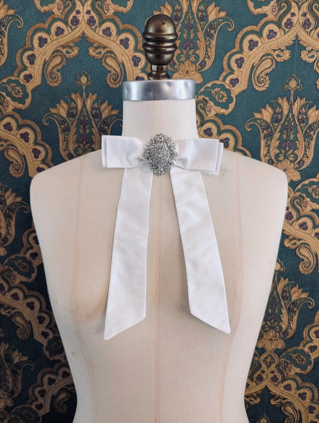 Broochella embellished bridal bow brooch in ivory with a embellished brooch centre. Pinned on a mannequin. The mannequin is dressed in a white, beaded, strapless wedding dress. The background is green and gold damask wallpaper.
