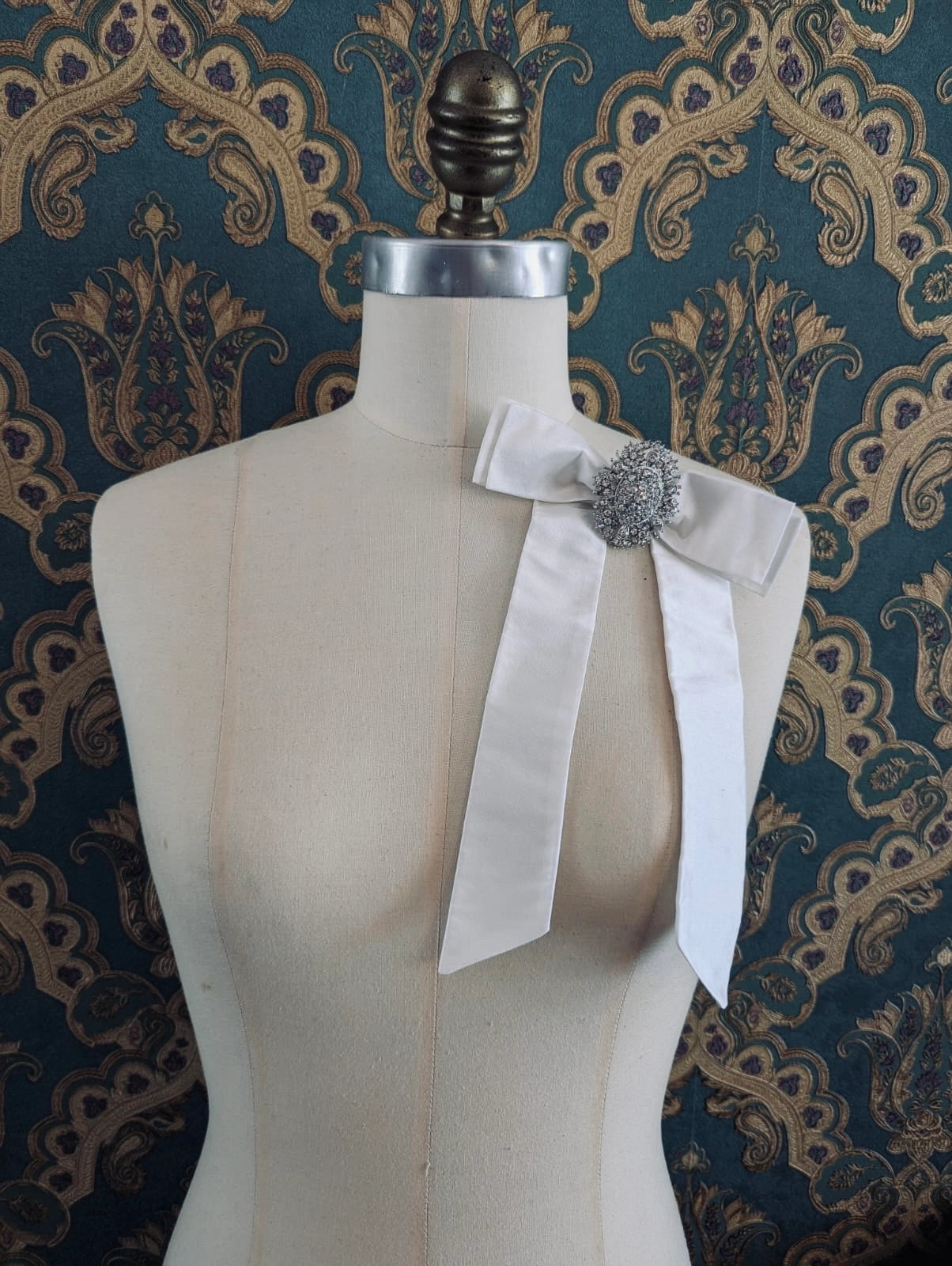 Broochella embellished bridal bow brooch in ivory with a embellished brooch centre. Pinned on a mannequin. The mannequin is dressed in a white, beaded, strapless wedding dress. The background is green and gold damask wallpaper.