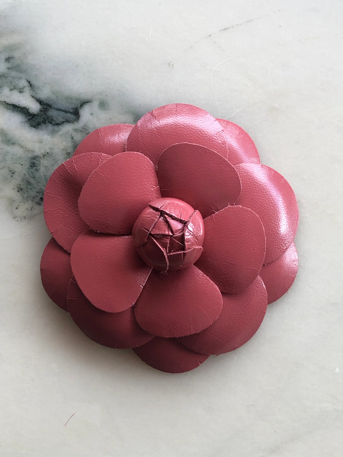 Broochella Camelia Leather Flower Brooch in coral pink, featuring handcrafted layered kid-skin leather petals and a sculptural flower centre on a cream marble background.