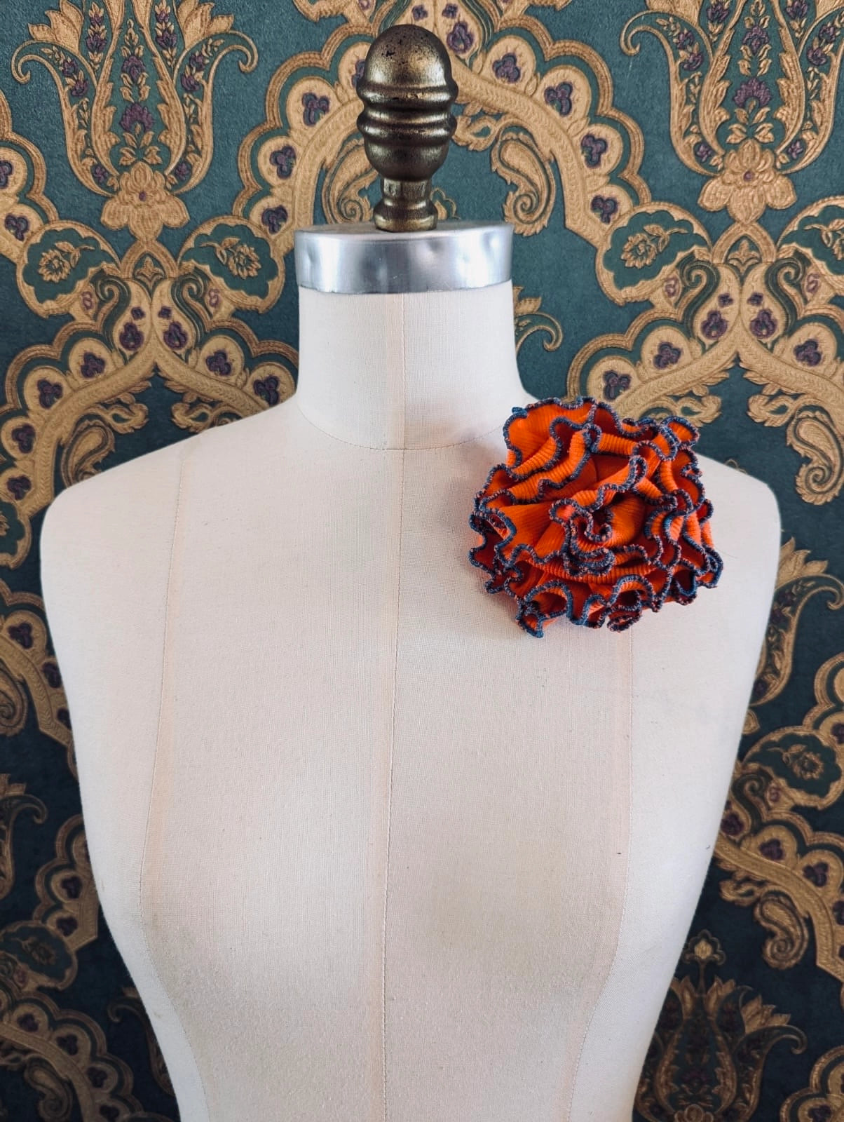 Broochella Corallo handmade fabric flower coral brooch. Made from orange stretch fabric, with a blue stitched edge. pinned to a mannequin. The background is green and gold damask wallpaper. 