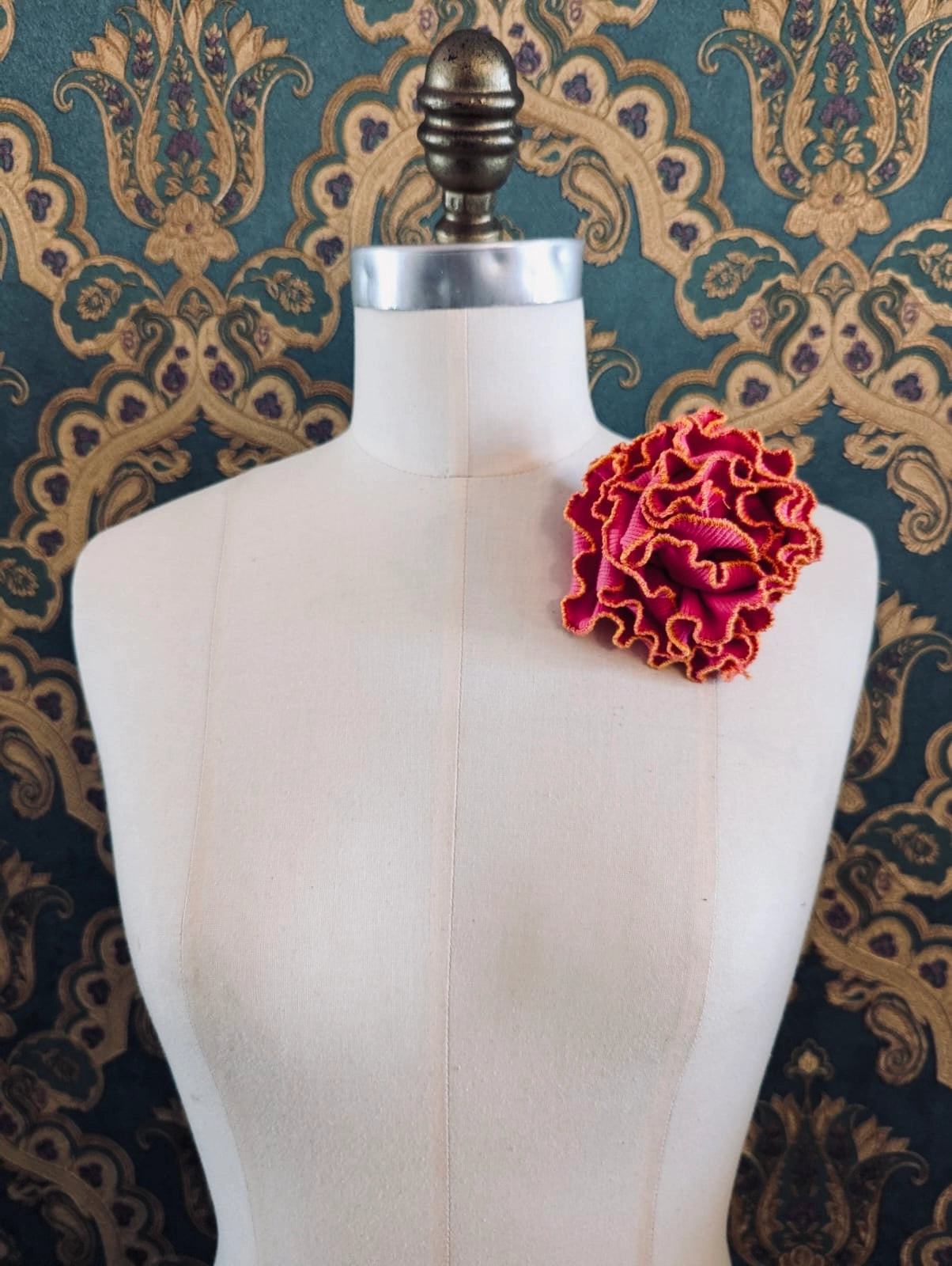 Broochella Corallo handmade fabric flower coral brooch. Made from pink stretch fabric, with a yellow and pink stitched edge. pinned to a mannequin. The background is green and gold damask wallpaper. 