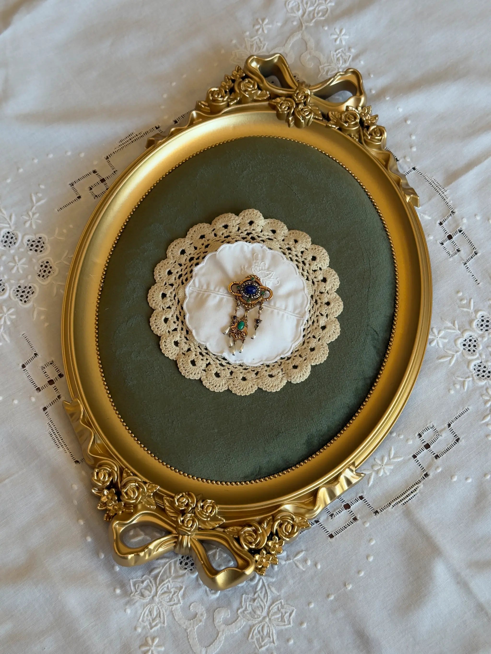 Broochella corredi 11/11 vintage, handmade linen, doily and lace brooch. The centre embellishment is a vintage gold tone metal and blue tone stone brooch. It is pictured on a green velvet tray with a gold frame. The background is a vintage, white linen tablecloth. 