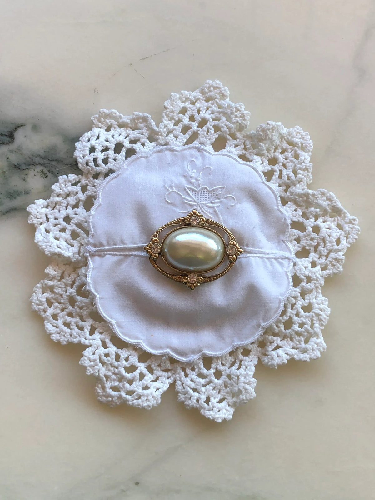 Broochella Corredi brooch pin handcrafted from a white vintage crochet doily and embroidered scalloped linen centre, finished with an ornate gold-tone vintage brooch featuring a large luminous pearl-style oval centre and delicate crystal accents, photographed on a white marble background with soft grey-green veining.