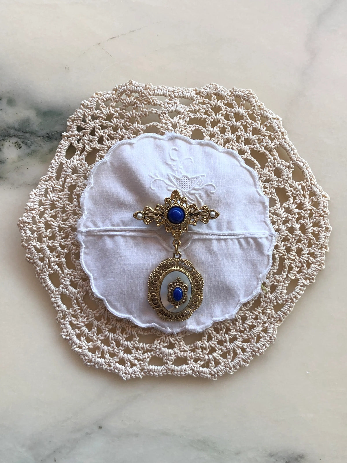 Broochella Corredi brooch pin handcrafted from an ivory vintage crochet doily and white embroidered scalloped linen centre, finished with an ornate gold-tone vintage brooch featuring deep blue stones, pearl accents and a decorative oval drop, photographed on a white marble background with soft grey-green veining.