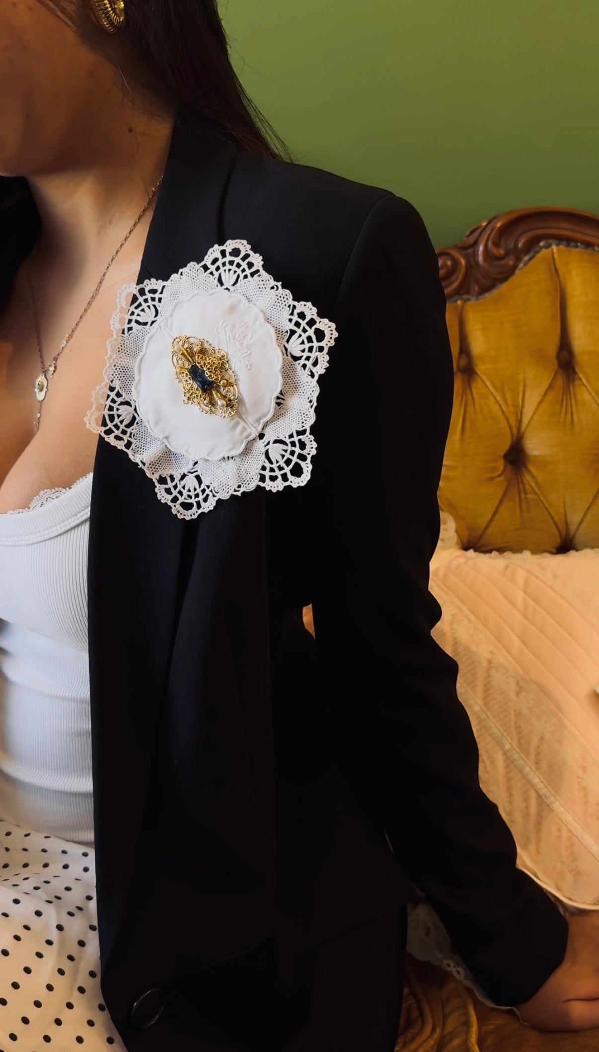 A close-up of a person wearing a black blazer with a large Broochella Corredi vinttage white lace floral brooch on the lapel, seated in front of a green wall and a tufted yellow chair.