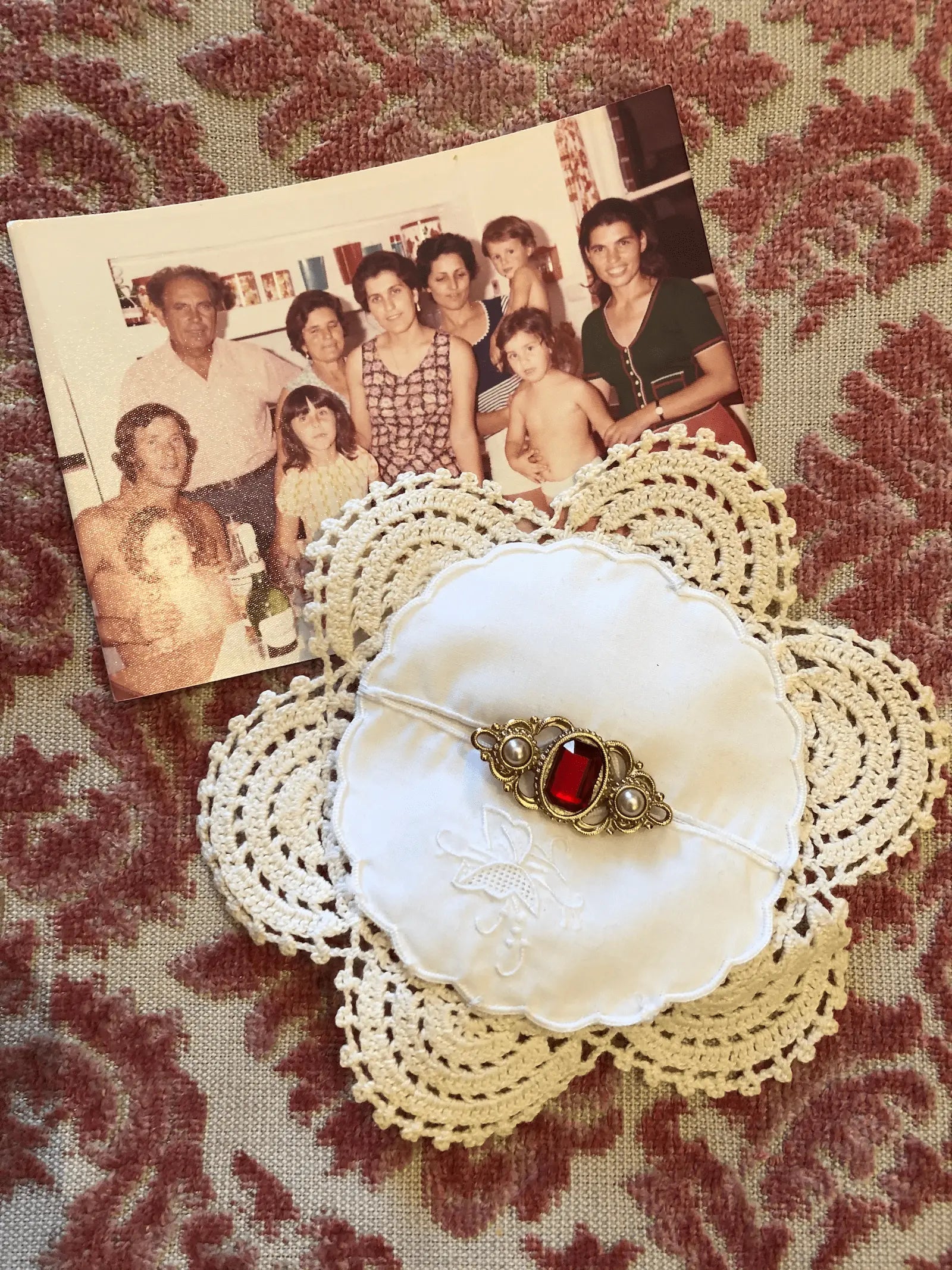 A Broochella Corredi 9/11 vintage doily, linen and lace brooch, with a red stone vintage brooch embellishment. An old family photograph is in the background on a patterned pink damask fabric surface.