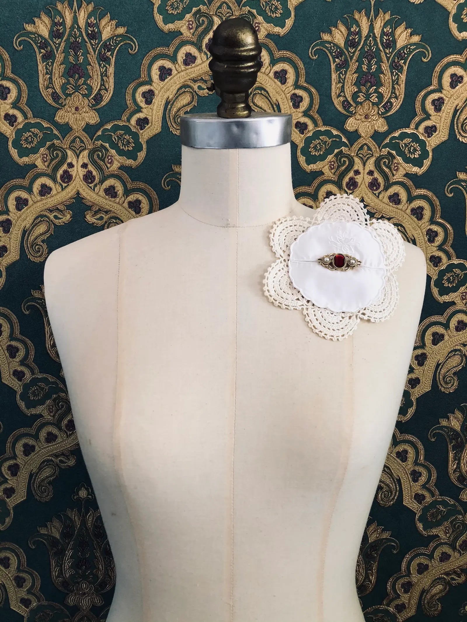 Broochella Corredi 9/11 handmade vintage lace, linen and doily brooch. The centre embellishment is a vintage gold tone metal brooch, wth a red stone centre. It is pinned to a mannequin. The background is green and gold damask wallpaper. 