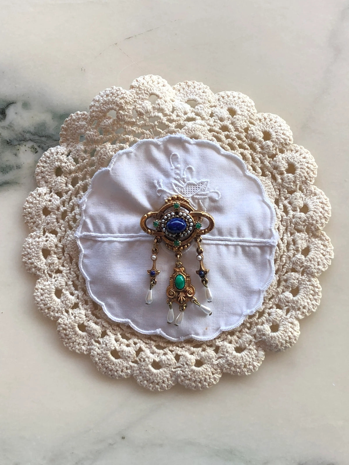Broochella Corredi brooch pin handcrafted from layered ivory crochet and white embroidered vintage doilies, centred with an ornate gold-tone vintage brooch featuring deep blue and turquoise stones, pearl accents and delicate dangling details, photographed on a white marble background with soft grey-green veining.