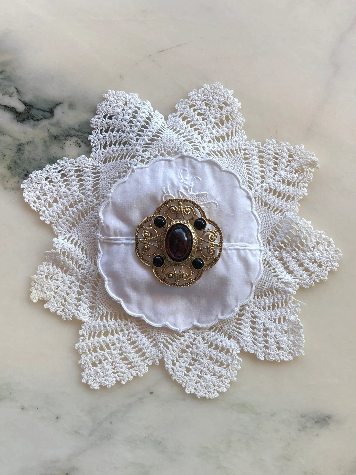 Broochella Corredi brooch pin handcrafted from a white vintage embroidered crochet doily, finished with an ornate gold-tone filigree vintage brooch featuring a deep burgundy oval centre stone and four black stones, photographed on a white marble background with soft grey-green veining.