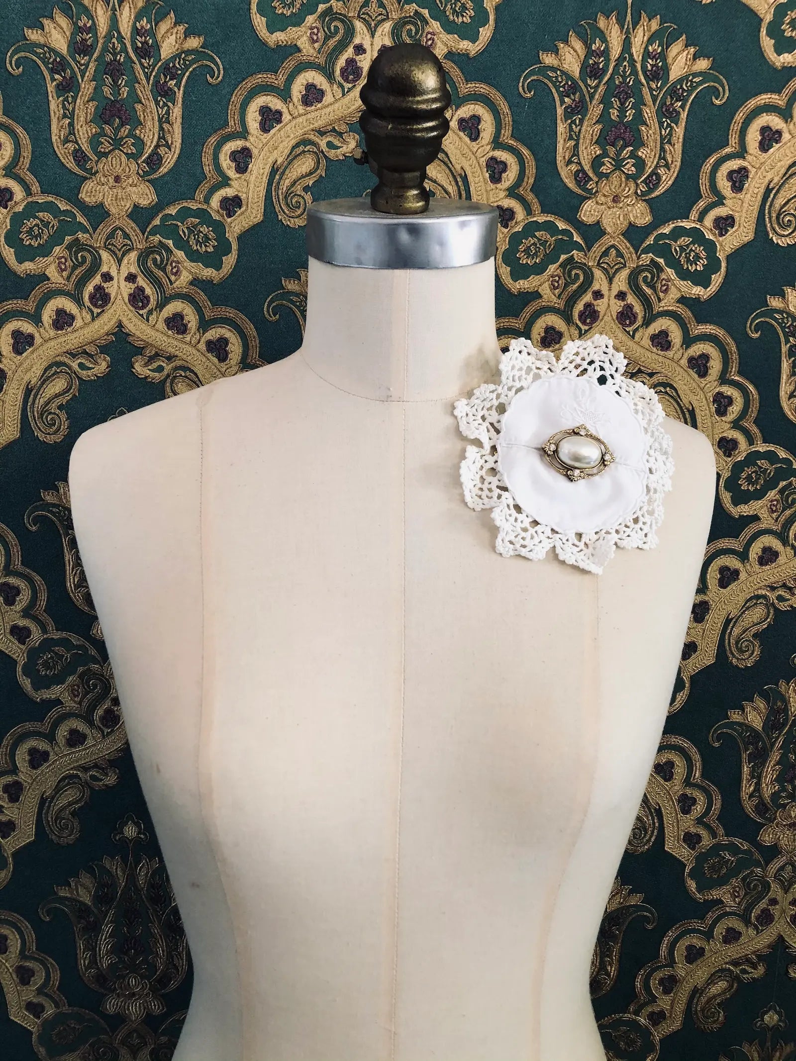 White Broochella Corredi 6/11 vintage lace, linen and doily handmade brooch. In the centre is a gold and pearl vintage brooch embellishment. It is pinned on a mannequin bust against a decorative wallpaper background