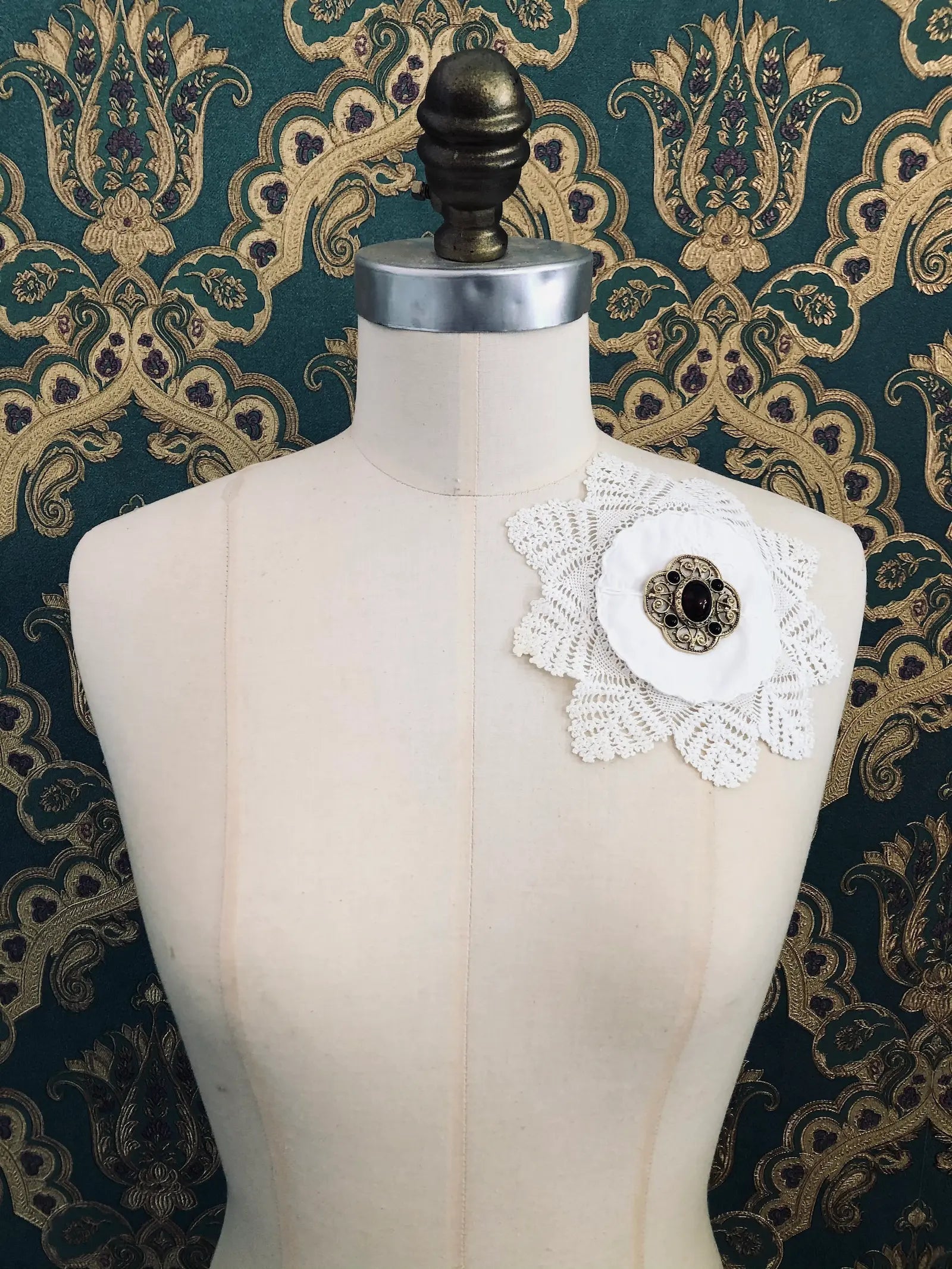 Broochella Corredi 4/11 vintage white doily and lace brooch with a vintage brooch embellishment. It is pinned to a mannequin. The background is green and gold damask wallpaper.