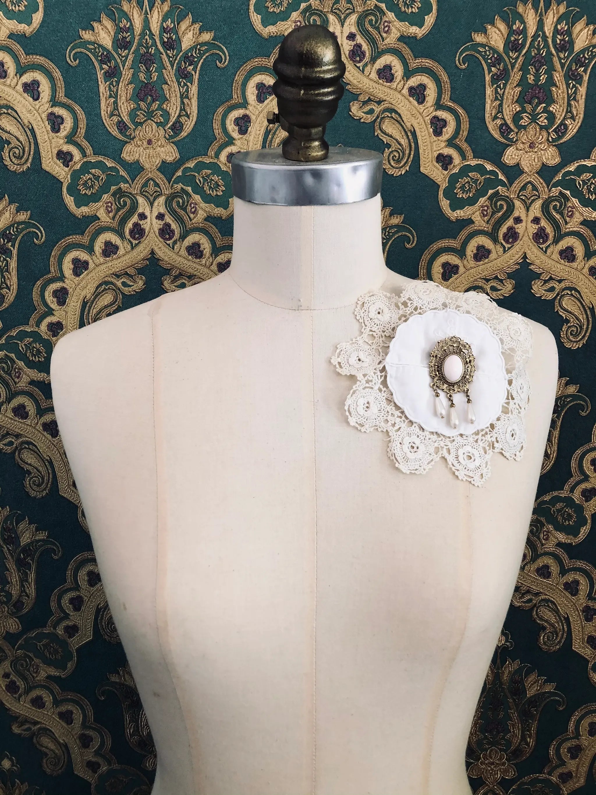 Broochella Corredi 1/11 vintage white doily and lace brooch with a vintage brooch embellishment. It is pinned to a mannequin. The background is green and gold damask wallpaper.