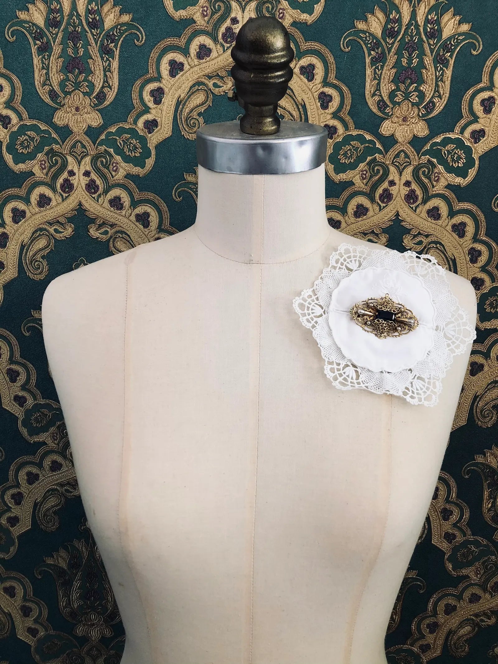 Broochella Corredi 5/11 vintage white doily and lace brooch with a vintage brooch embellishment. It is pinned to a mannequin. The background is green and gold damask wallpaper.