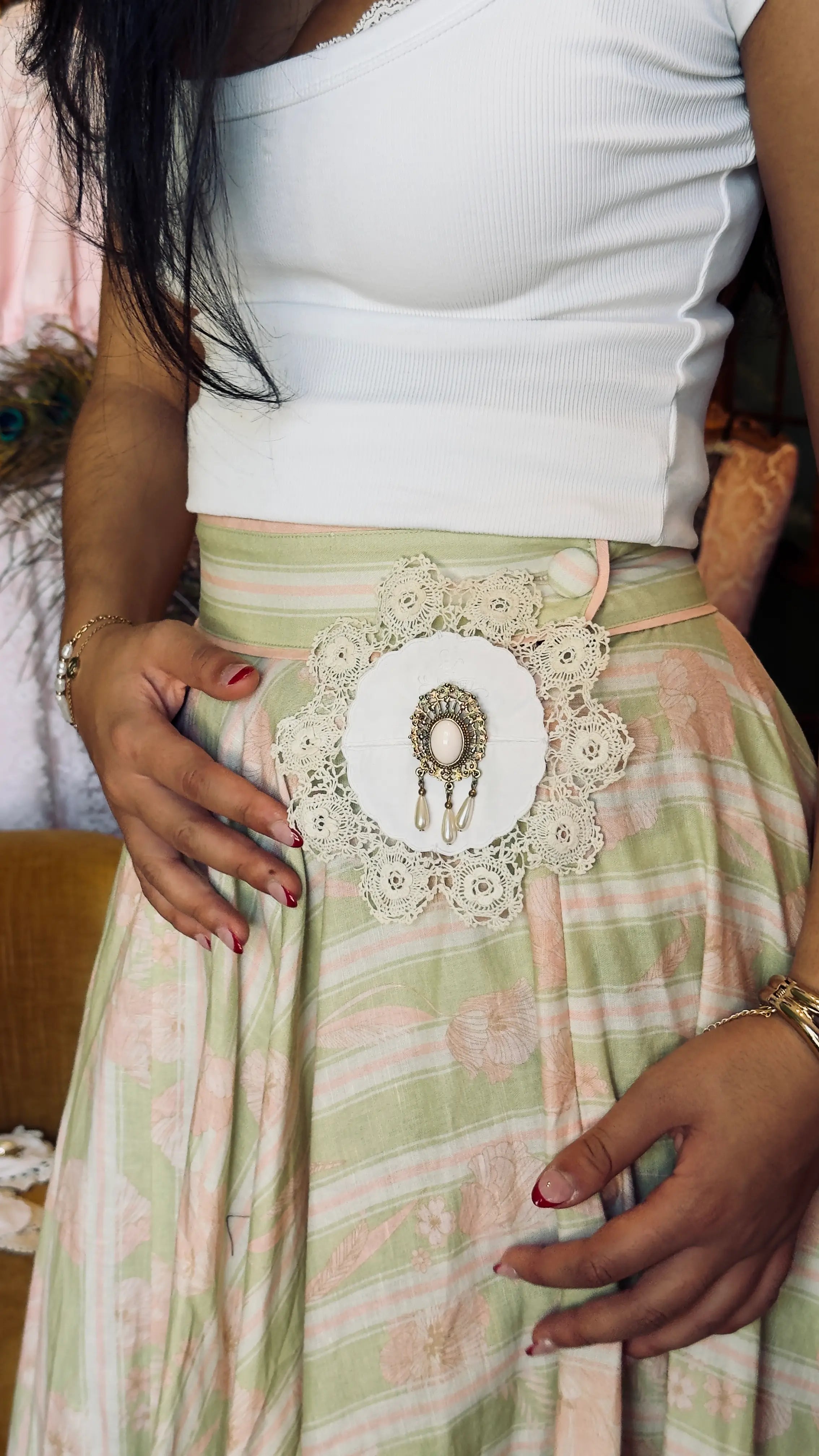 A close-up of a person wearing a white ribbed tank top and a light green and pink striped skirt with a Broochella Corredi brooch 1/11. It has a large white lace applique and central decorative patch at the waist.