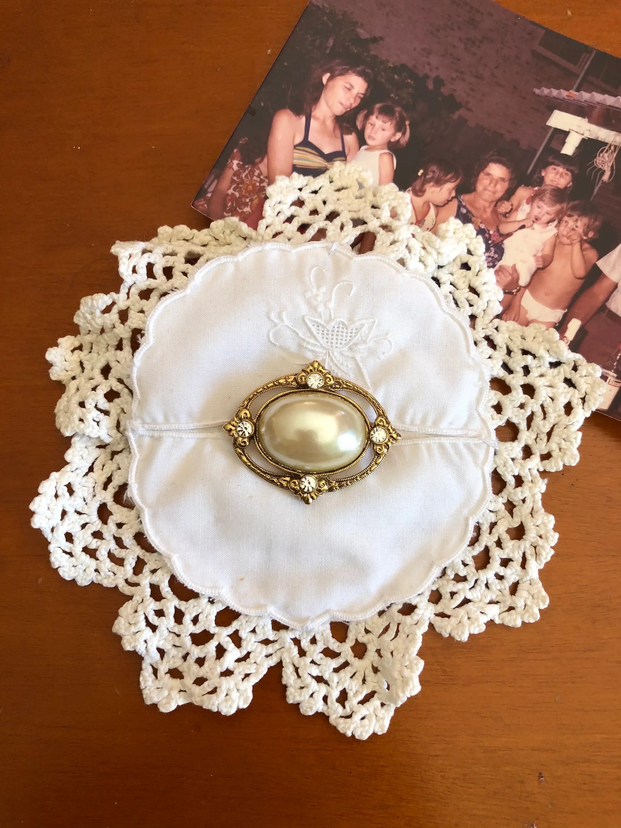 A  Broochella Corredi 6/11 vintage brooch featuring a large cream-coloured pearl or glass bead surrounded by gold-toned metal and small clear stones, displayed on a white embroidered fabric circle over a cream lace doily on a wooden surface, with an old family photograph in the background.
