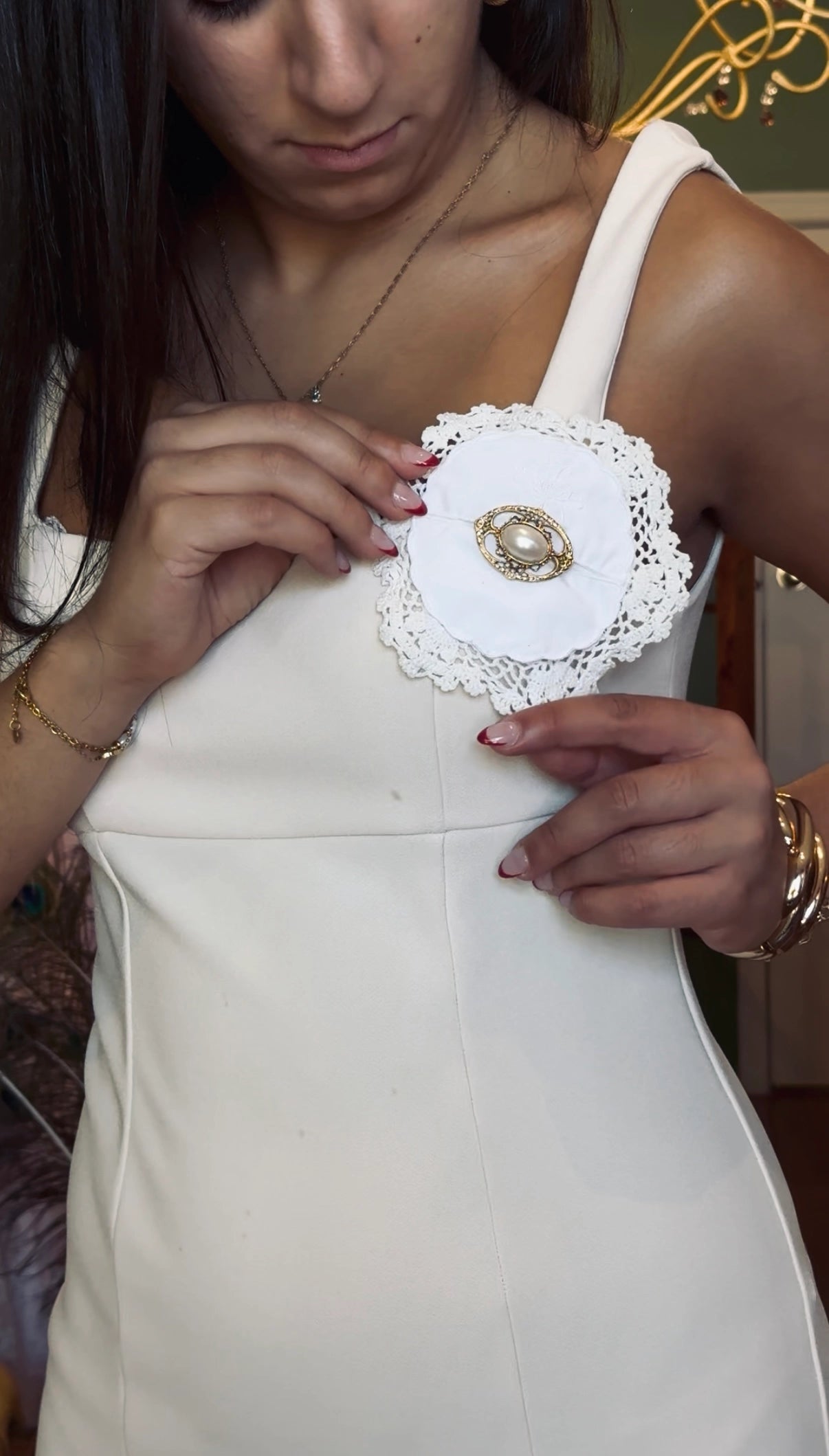 A close-up of a woman wearing a white sleeveless dress and adjusting a Broochella Corredi white lace applique brooch, with a gold and pearl vintage brooch embellishment with a gold circular centre on the bodice.