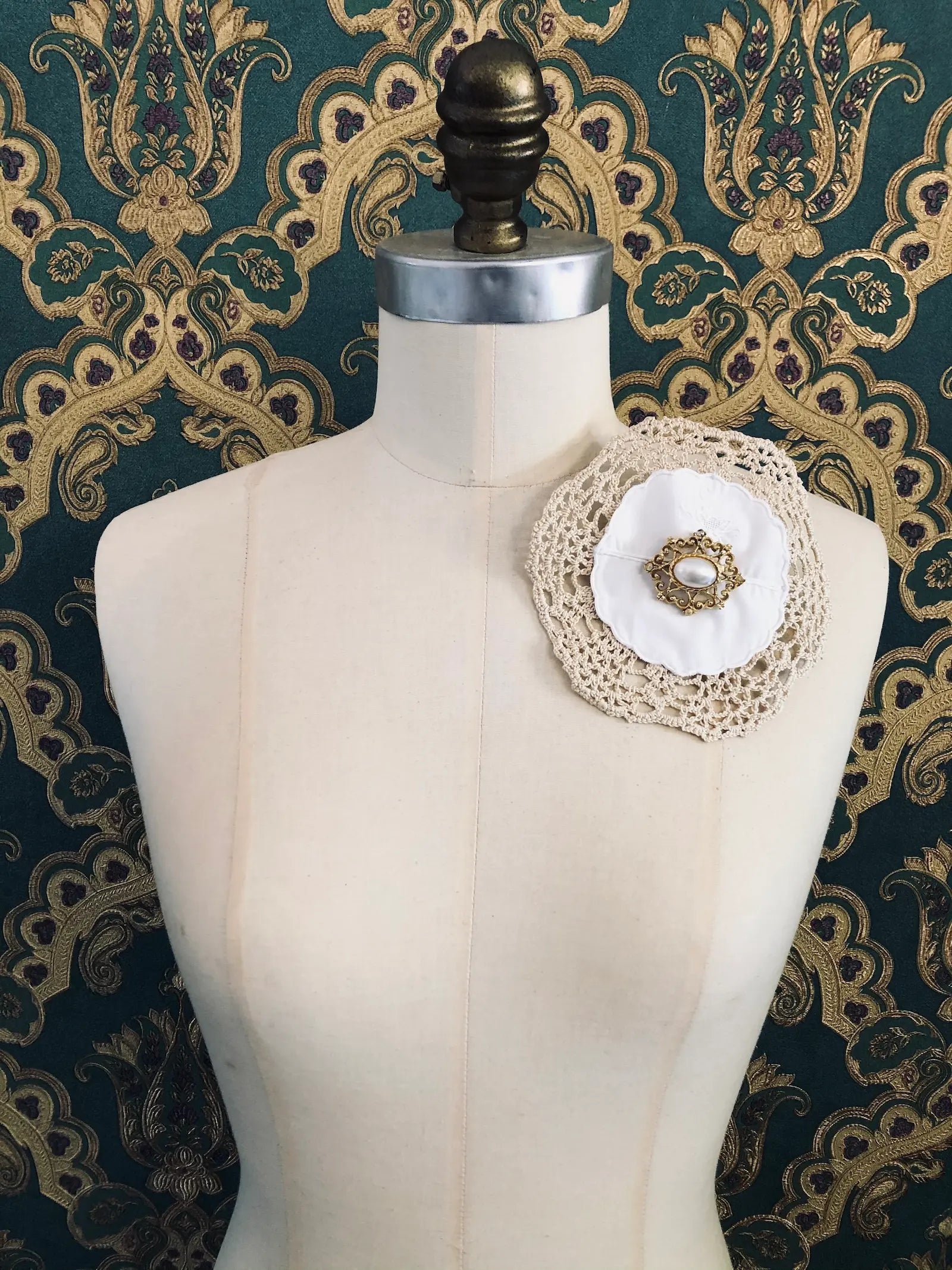 Broochella Corredi 10/11 handmade vintage lace, linen and doily brooch. The centre embellishment is a vintage gold tone metal brooch, wth a pearl centre. It is pinned to a mannequin. The background is green and gold damask wallpaper. 