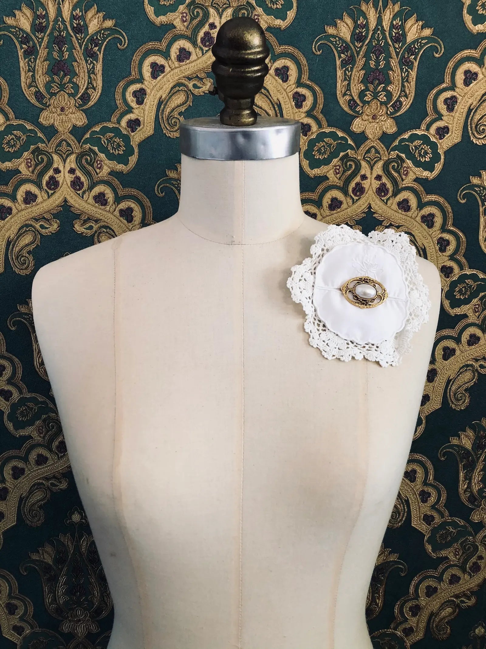A Broochella Corredi 7-11 white fabric, vintage linen and lace doily brooch with a central gold-toned pin and white lace trim, displayed on a cream-coloured dress form against a dark green and gold damask patterned backdrop.